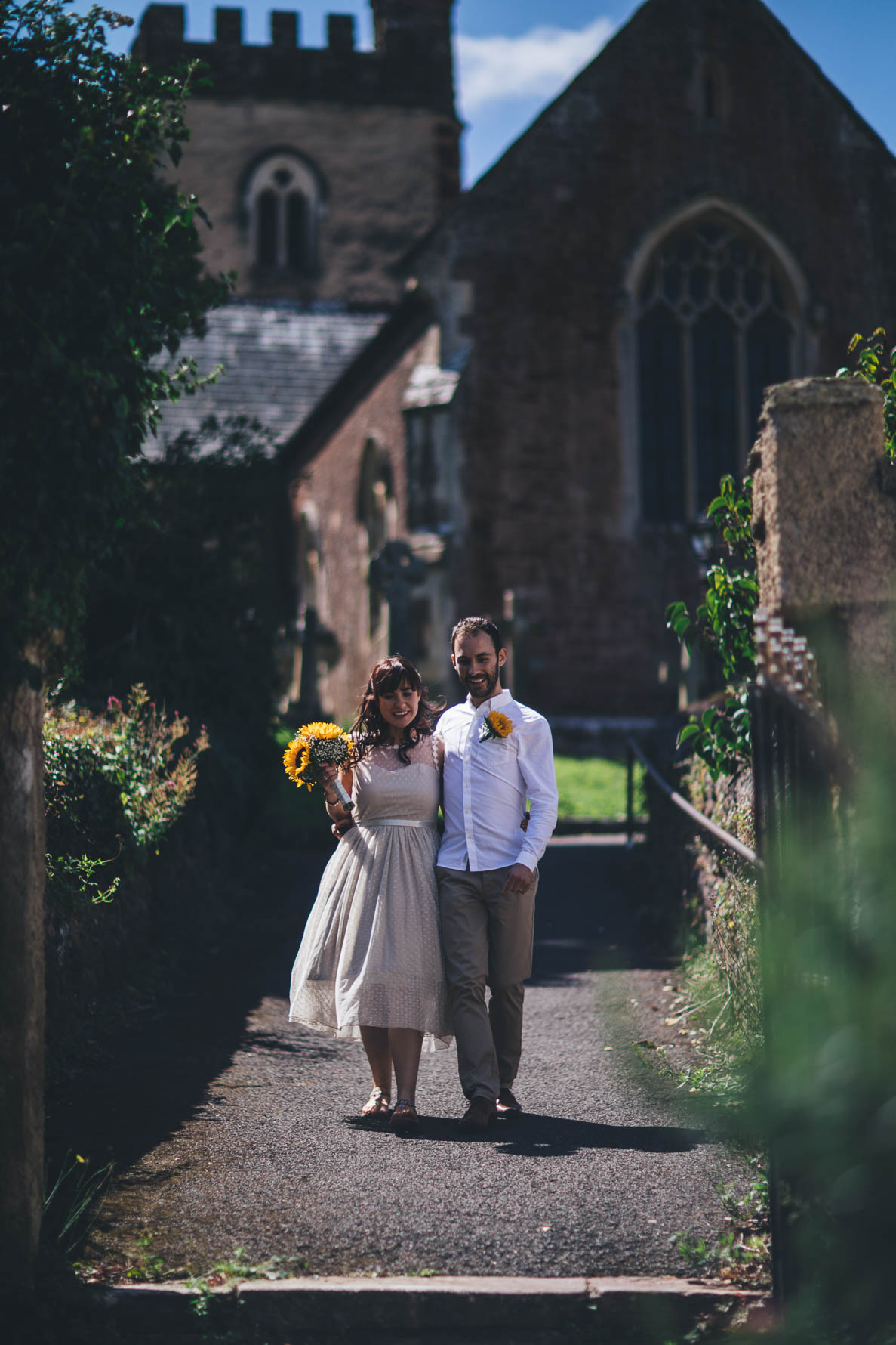 Bride and groom walking down a path in a churchyard away from the church. The groom has his arm around the bride's waist. The bride is holding a bouquet of sunflowers and the groom has a sunflower pinned to his white shirt.