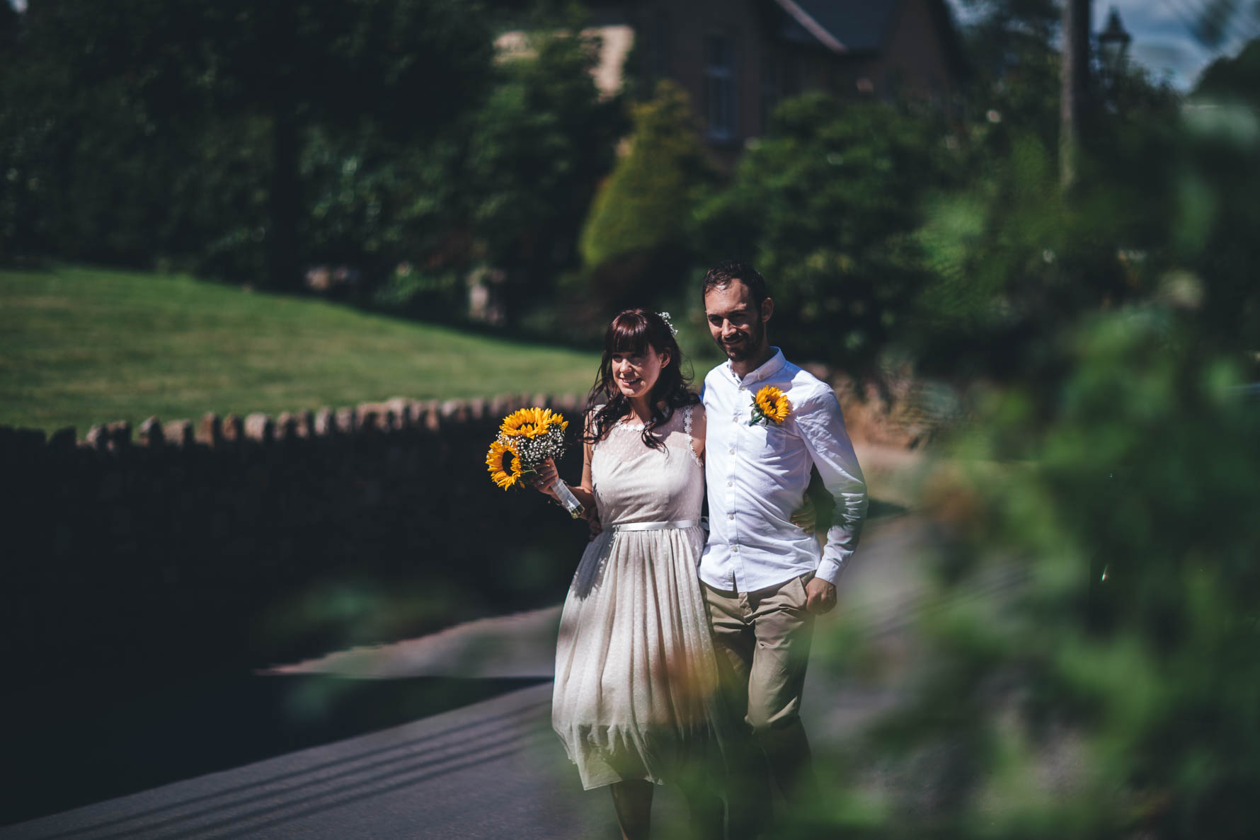 Bride and groom walking down a path in a churchyard. The groom has his arm around the bride's waist. The bride is holding a bouquet of sunflowers and the groom has a sunflower pinned to his white shirt.