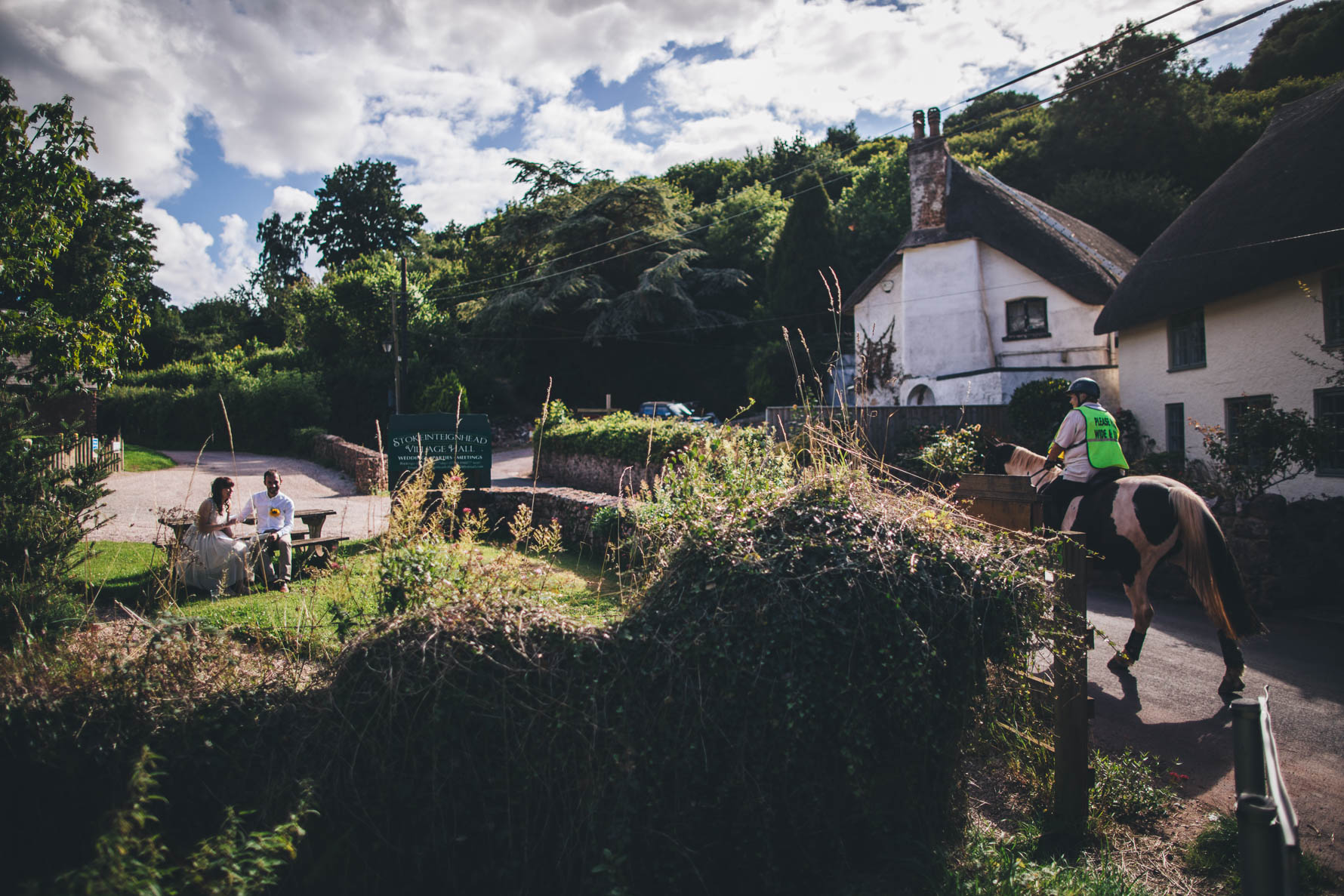 Bride and groom sat to the left of the picture on a bench as a person is about to ride by on a horse to the right of the pictur in front of some white-washed thatched cottages
