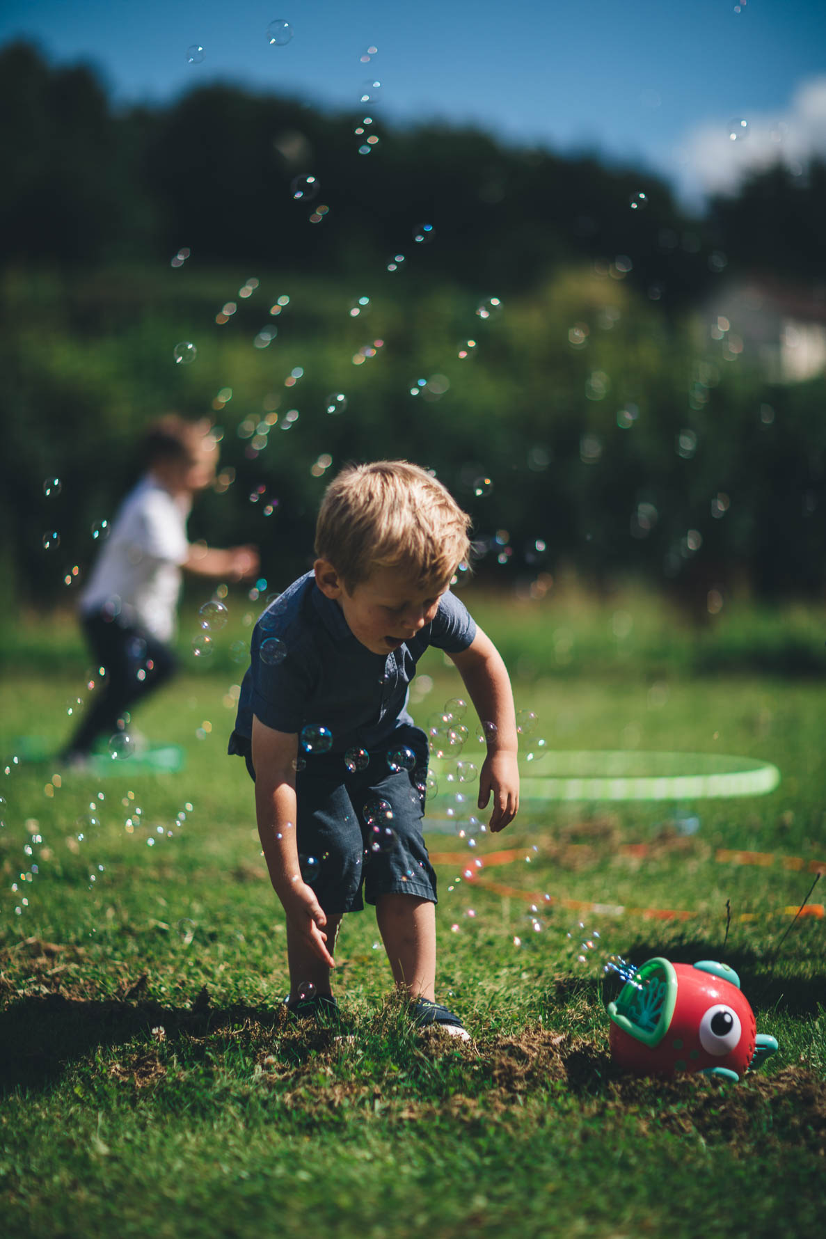Young boy stood on a lawn bending down to catch some bubbles which are being blown out of a fish-shaped bubble machine