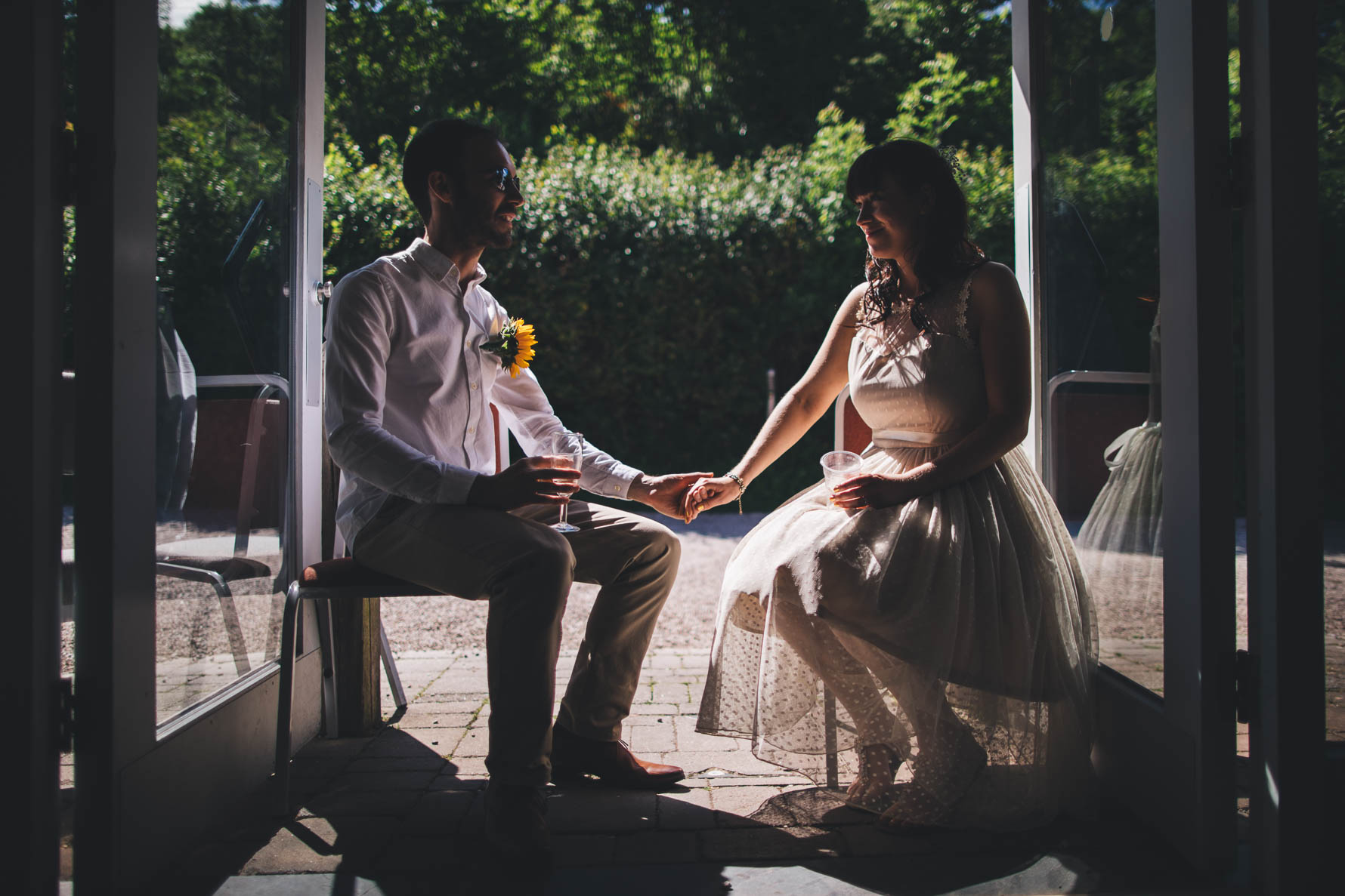 Bride and groom sat in a doorway of a village hall in Devon holding hands. Both are holding drinks and the groom is wearing sunglasses and has a sunflower pinned to his white shirt