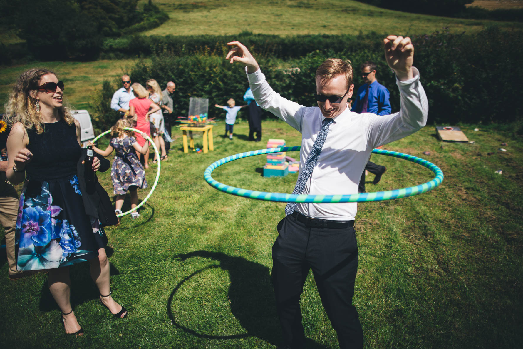 Man wearing a shirt and tie and sunglasses stood on a lawn rotating a hula hoop around his chest