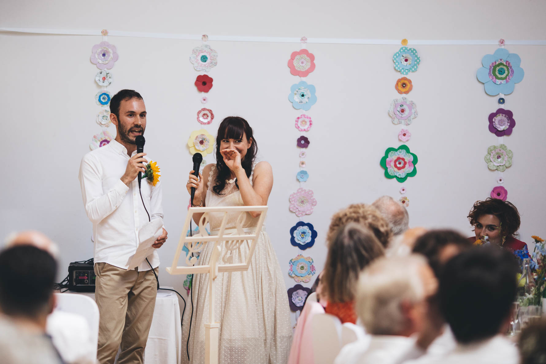 Bride and groom stood at the front of a village hall reading a speech during their wedding reception. The groom is talking into a microphone and the bride is holding a microphone but has her other hand to her mouth and is smiling. There are colourful paper flowers on the white wall behind them