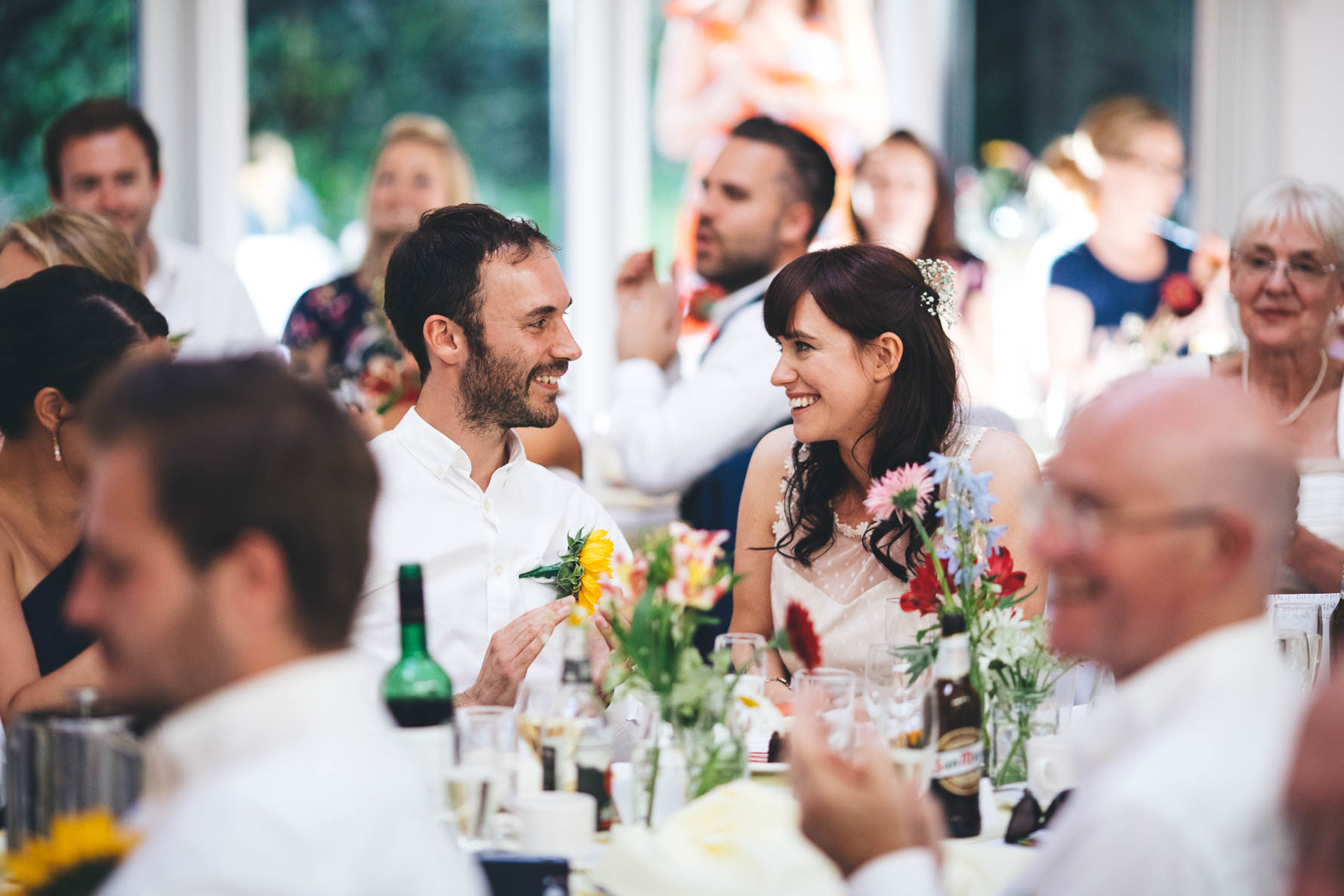 Bride and groom seated at their wedding reception smiling at each other as someone out of shot is reading a speech. There are other wedding guests around them who are out of focus