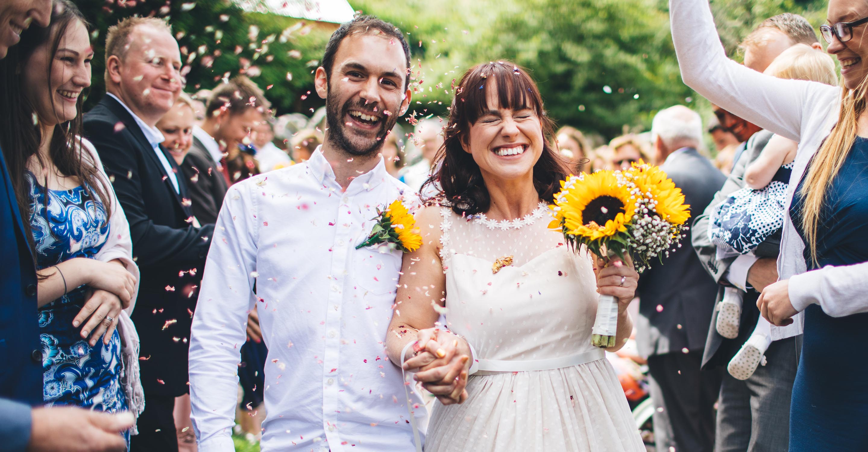 Bride and groom having confetti thrown over them. They are both smiling and the bride has her eyes scrunched up and closed. The bride is holding a bouquet of sunflowers and the groom has a sunflower pinned to his white shirt