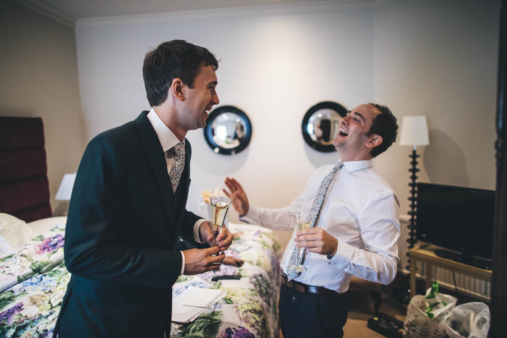 Groom and friend facing each other, laughing, next to a bed in a hotel room holding glasses of champagne.