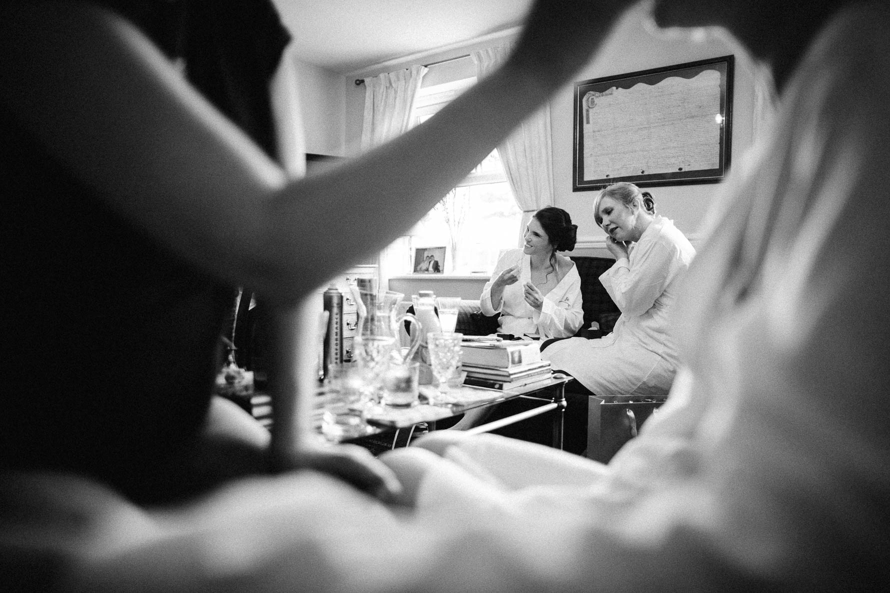 Black and white photo of bridesmaids wearing dressing gowns getting ready in the background while the bride is having her makeup done in the foreground