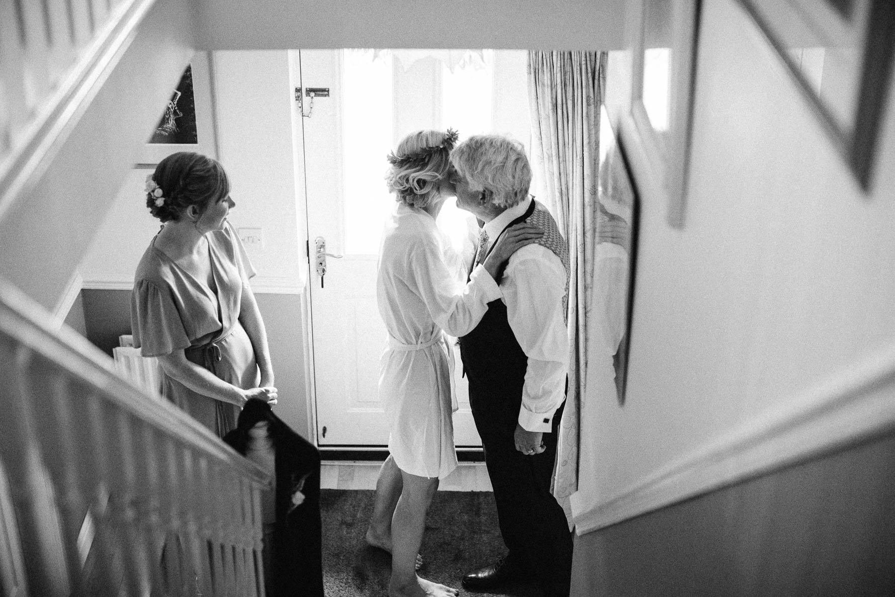 Black and white photo take from the top of the stairs of the bride, wearing a dressing gown, kissing her father at the foot of the stairs