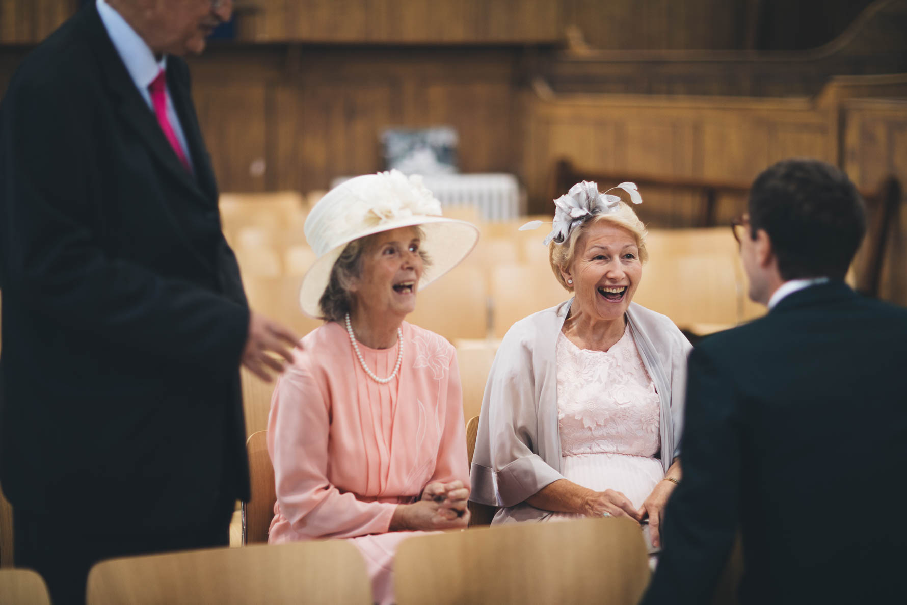 Two older ladies, one wearing a hat, the other a fascinator, seated, lauging and smiling looking towards the groom who is stood in front of them