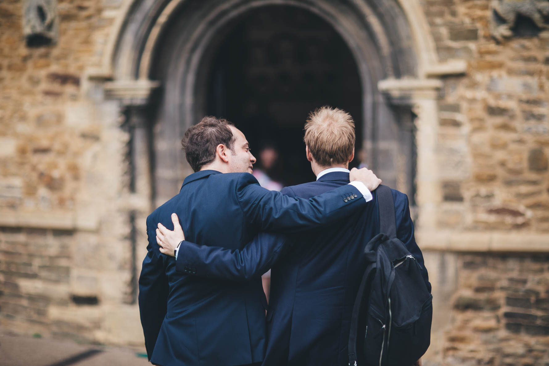 Picture taken from behind of two men wearing dark blue suits walking into Oakham Castle Great Hall with their arms around eah others shoulders. The man on the right has a black backpack over his shoulder
