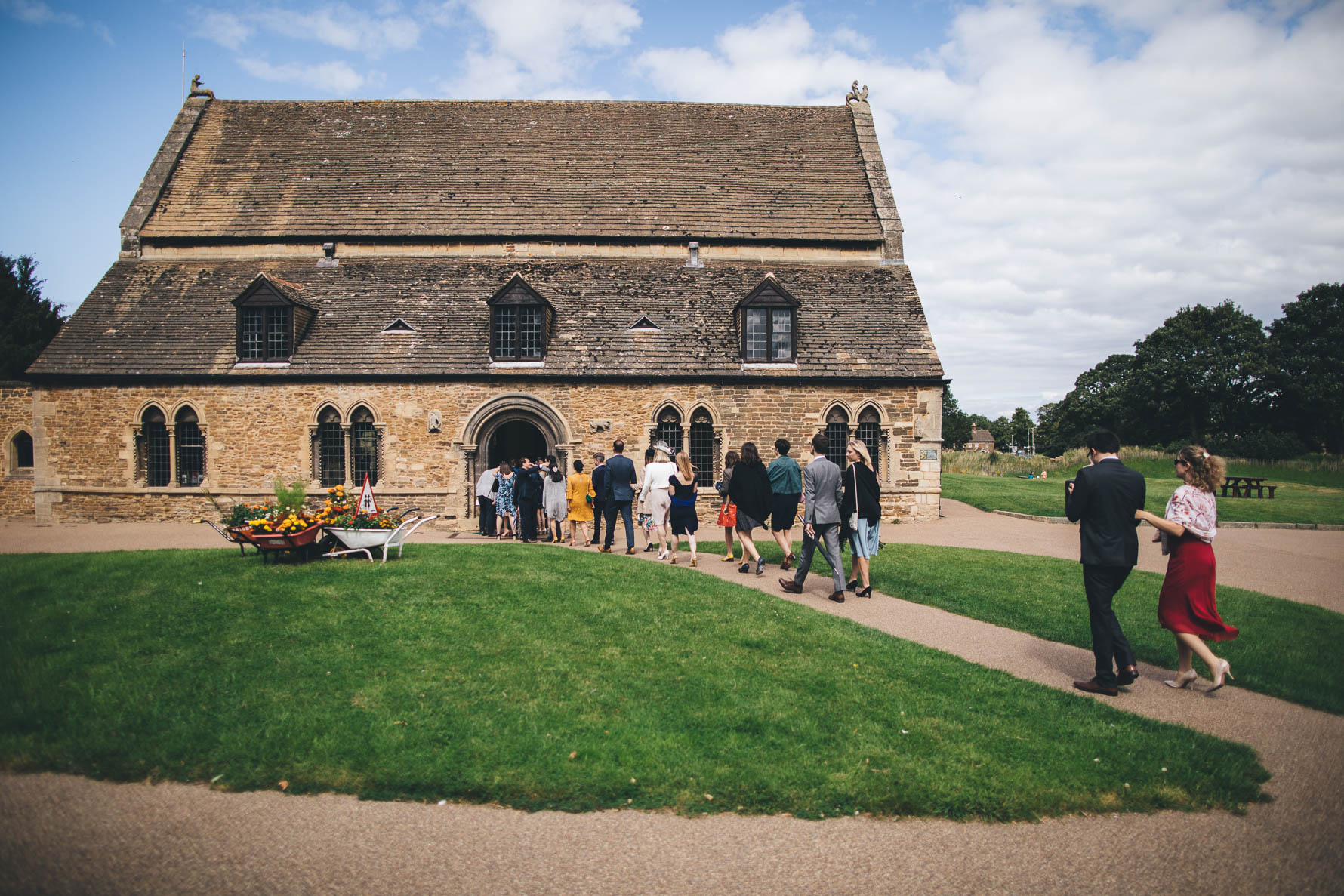 View of Oakham Castle Great Hall from outside with a lawned area to the front and right side and wedding guests walking up the path towards the entrance