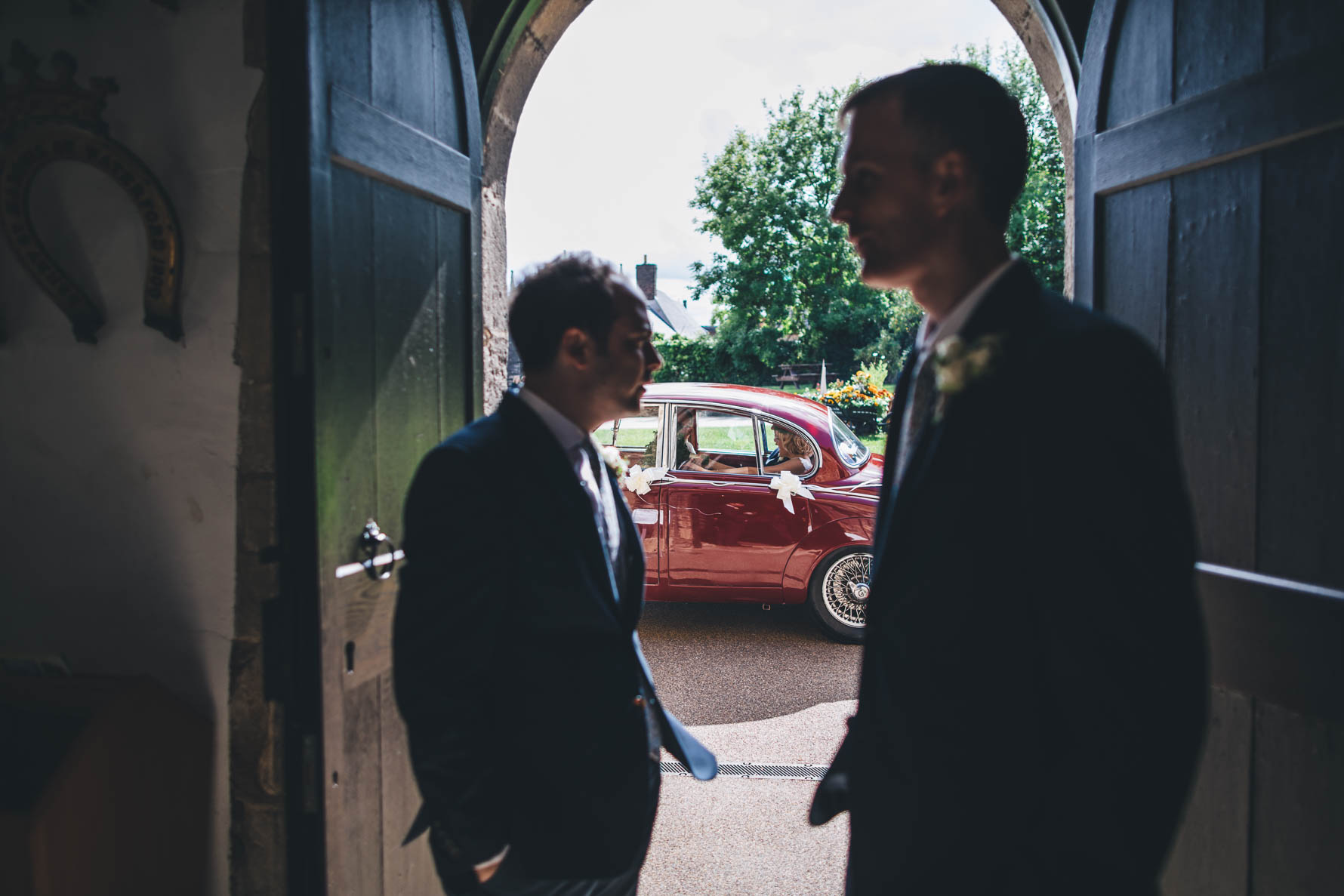 Two groomsmen waiting at the entrance of the Great Hall at Okham Castle as the bride pulls up in a vintage red car along with her father