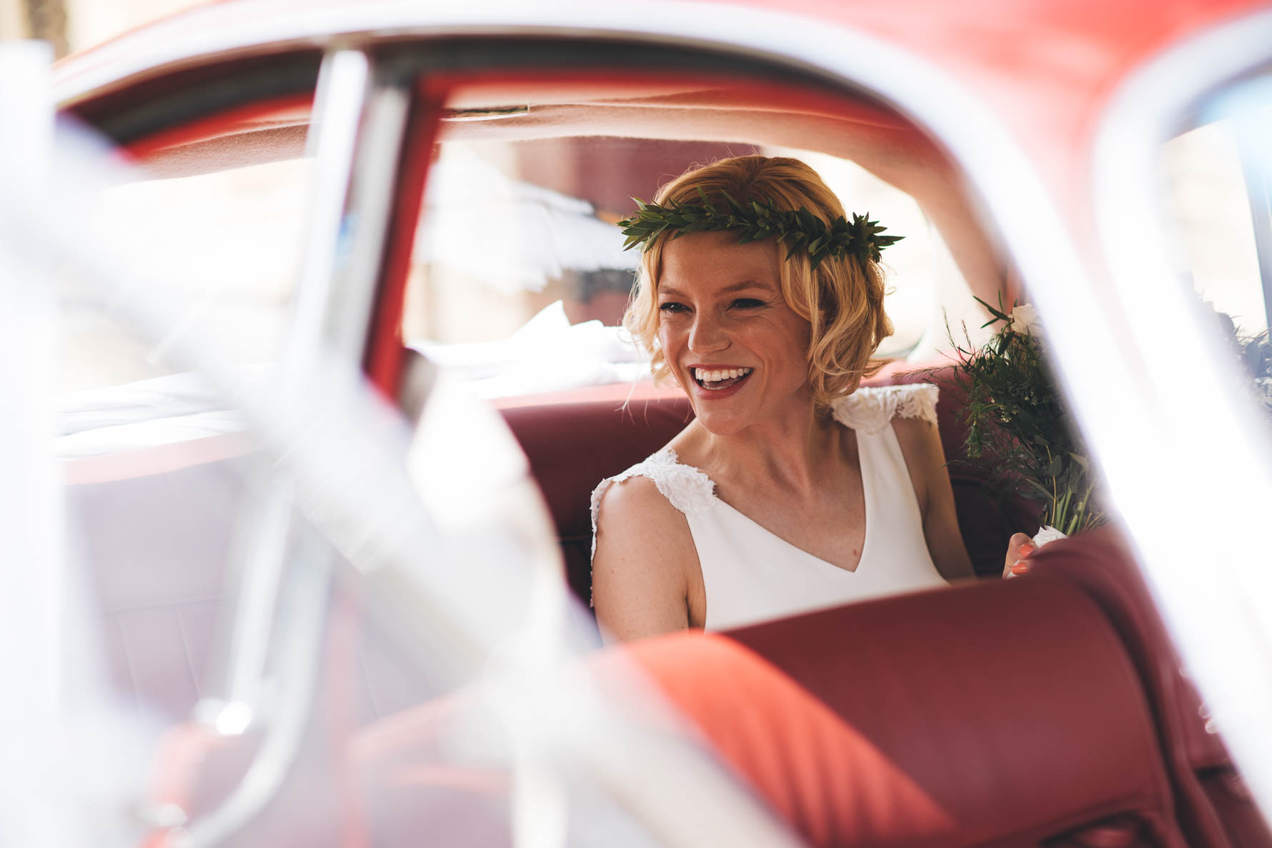 View through the open door of a vintage red car of the bride inside who has a leaf garland on her head