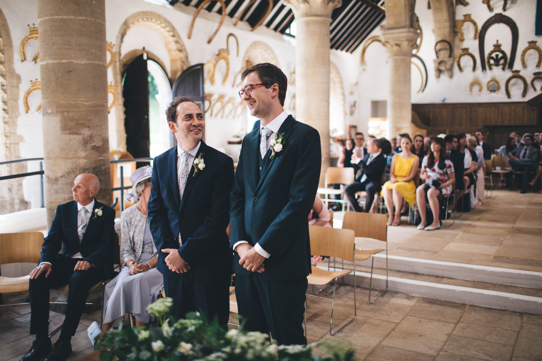 Groom and his best man waiting at the front of the Great Hall at Oakham Castle. There are ornamental horse shoes covering the walls