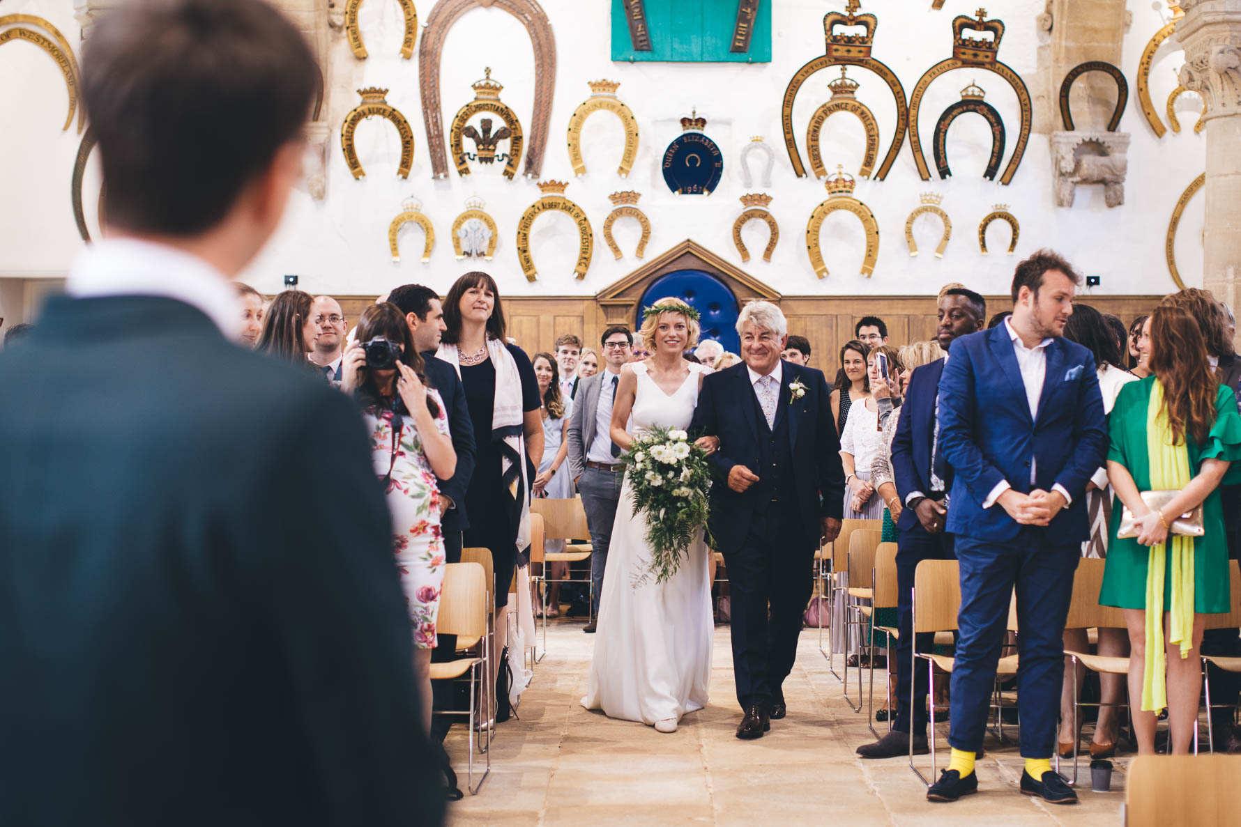 Bride being walked down the aisle by her father at the Great Hall at Oakham Castle. There are large ornamnetal horse shoes decorating the wall