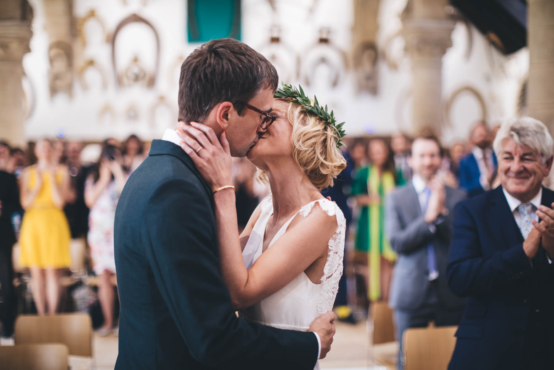 Bride and groom having their first kiss after getting married at the Great Hall at Oakham Castle with the wedding party in the background applauding