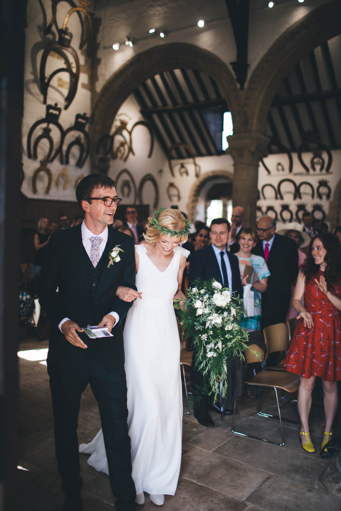 Bride and groom walking out of the Great Hall at Oakham Castle after getting married. The bride is holding a large bouquet of white floers with lots of green foliage. The wedding party can be seen stood in the background