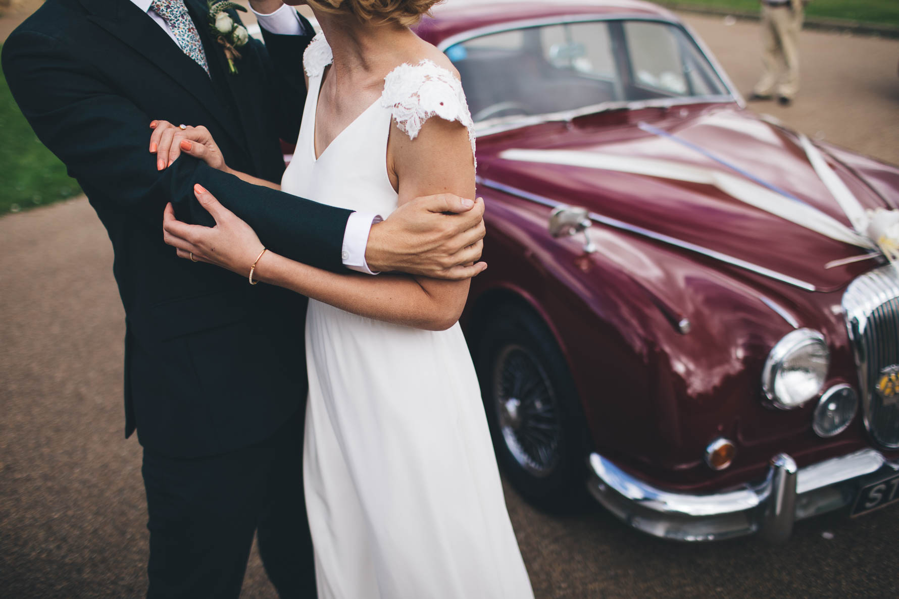 Bride and groom with their faces out of shot with their arms intertwined stood in front of a red vintage car