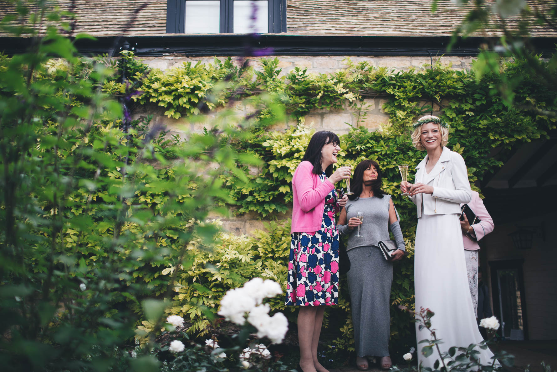 Bride wearing a white jacket stood outside a stone building covered in ivy along with three female wedding guests also holding glasses of champagne