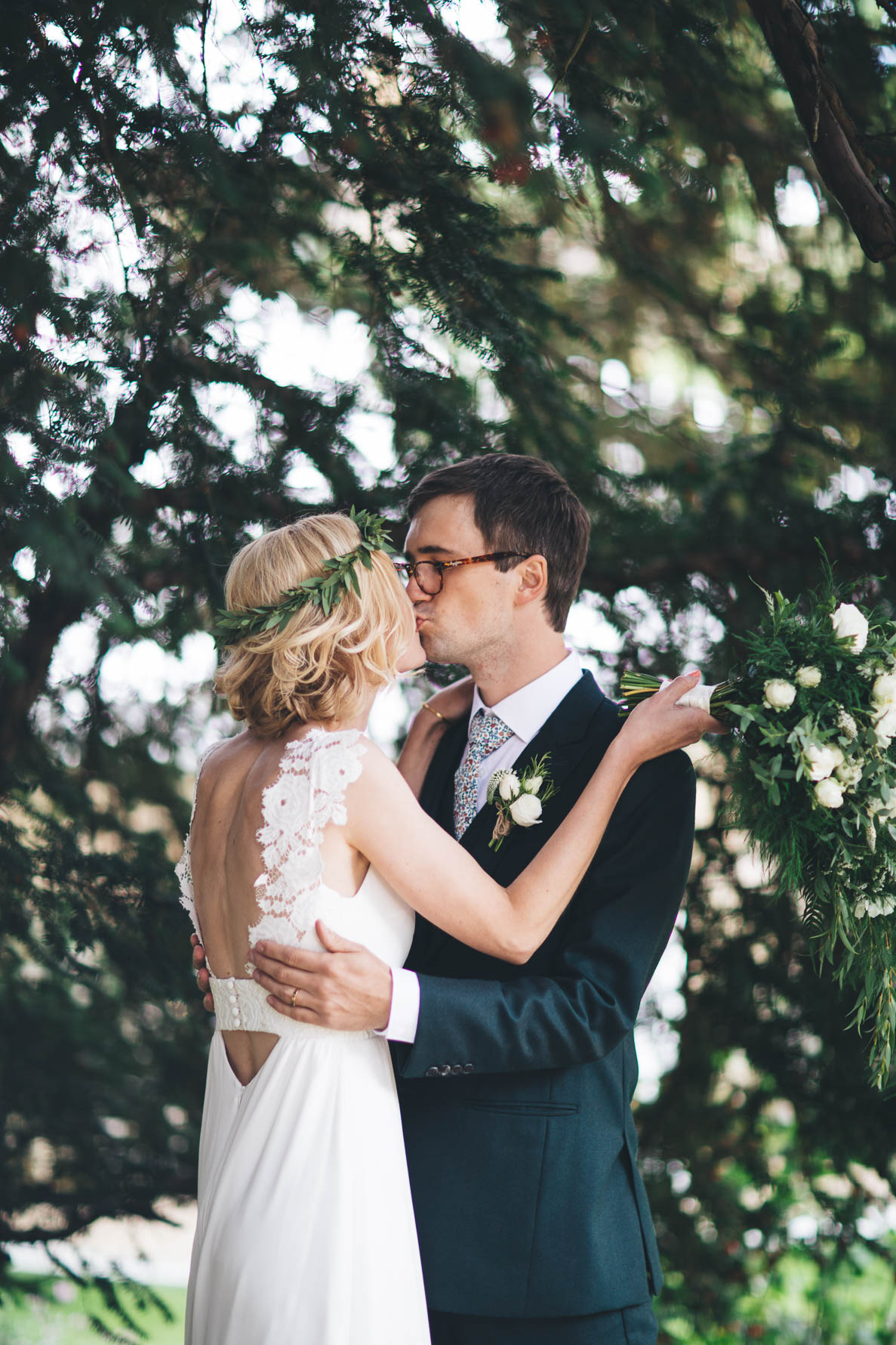 Bride and groom with their arms around each other stood sharing a kiss in a wooded area