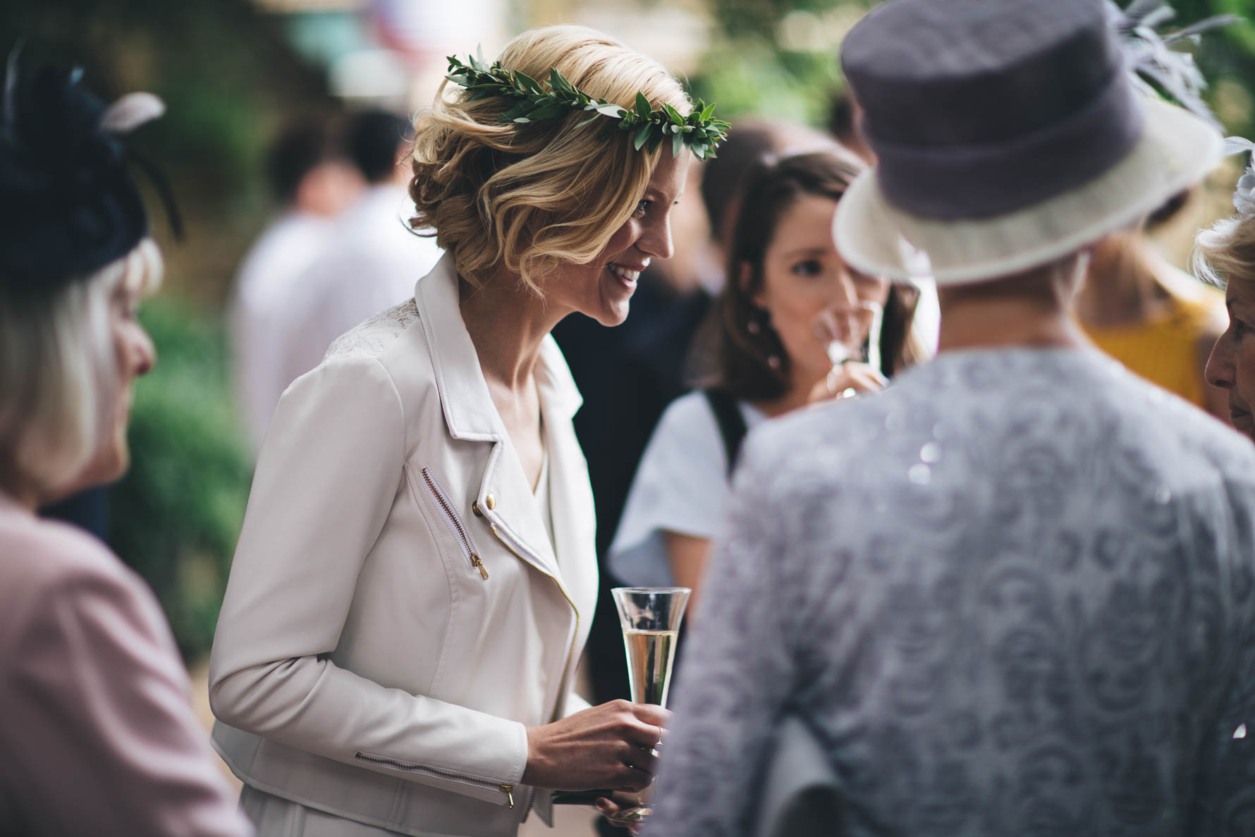 Bride wearing a white leather jacket and a leaf garland in her hair, holding a glass of champagne chatting to some female wedding guests outside