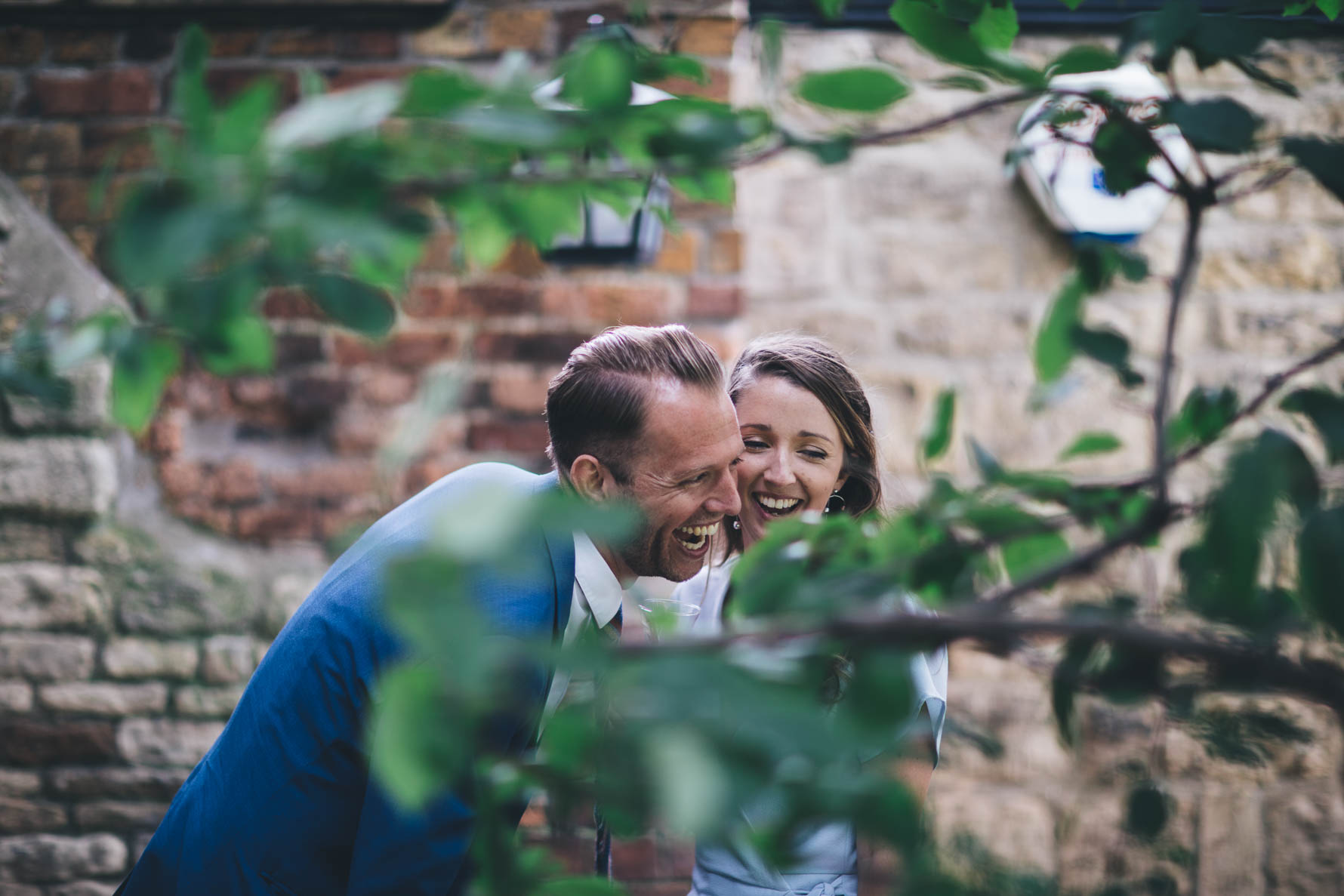 Shot taken through some branches of a tree of a couple laughing in front of a brick wall