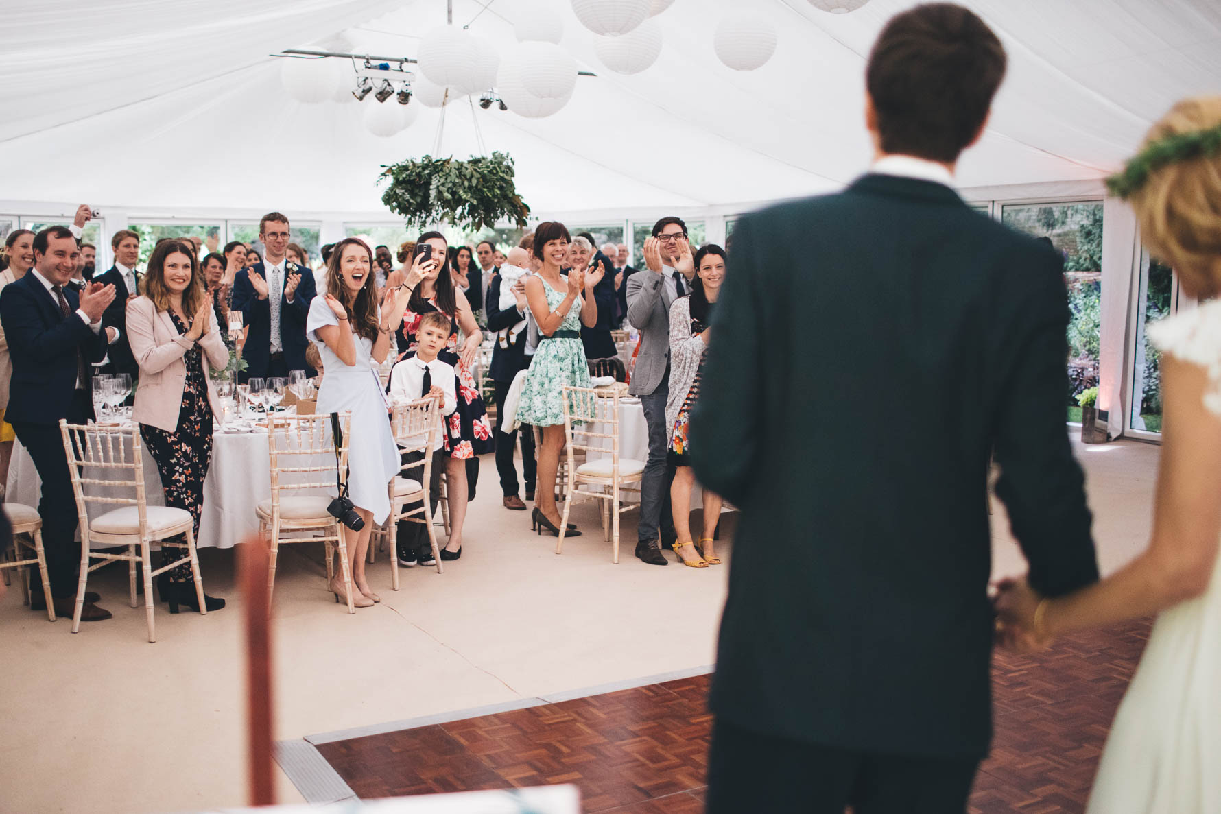 Bride and Groom, shot from behind, stood at the front of a marquee in the walled garden at the George of Stamford Hotel whilst the wedding guests are stood applauding their arrival in the background