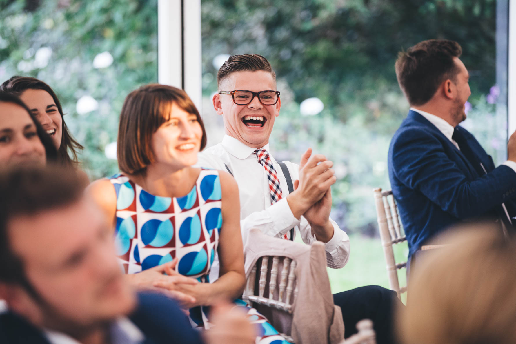Make wedding guest wearing glasses sat clapping with a large smile on his face surrounded by other wedding guests also smiling