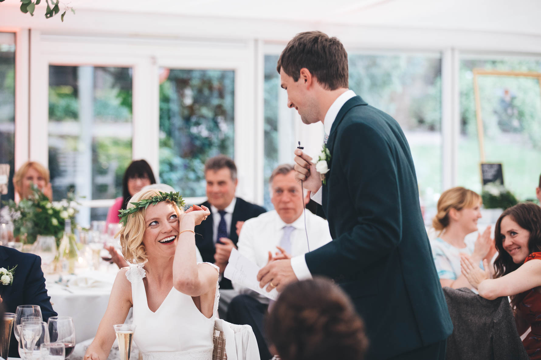 Groom stood in a marquee reading a speech as the bride is seated, wiping a tear away from her eye with members of the wedding party applauding in the background