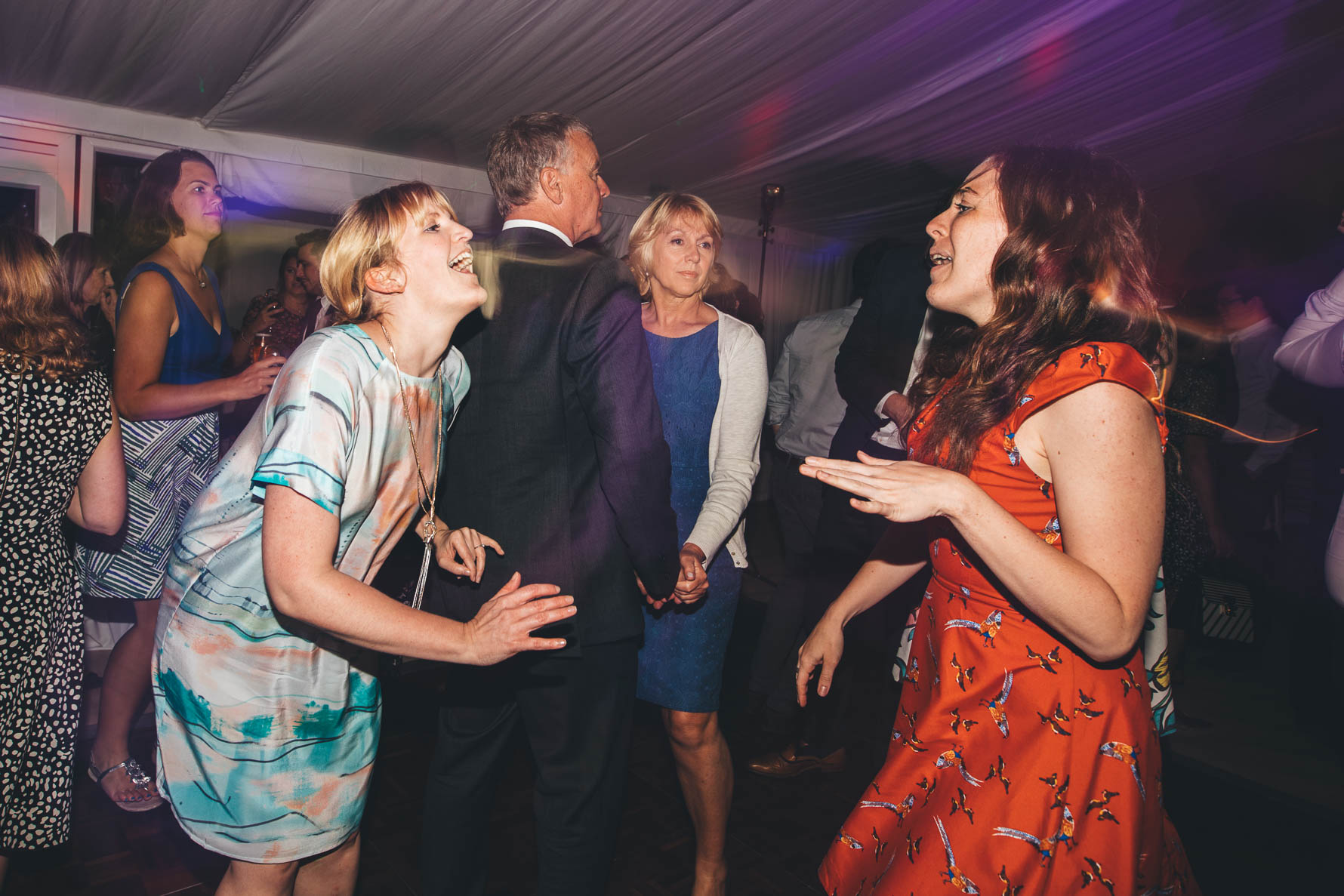 Two women facing each other on a dancefloor in a marquee singing along to a song as other members of the wedding party are dancing behind them