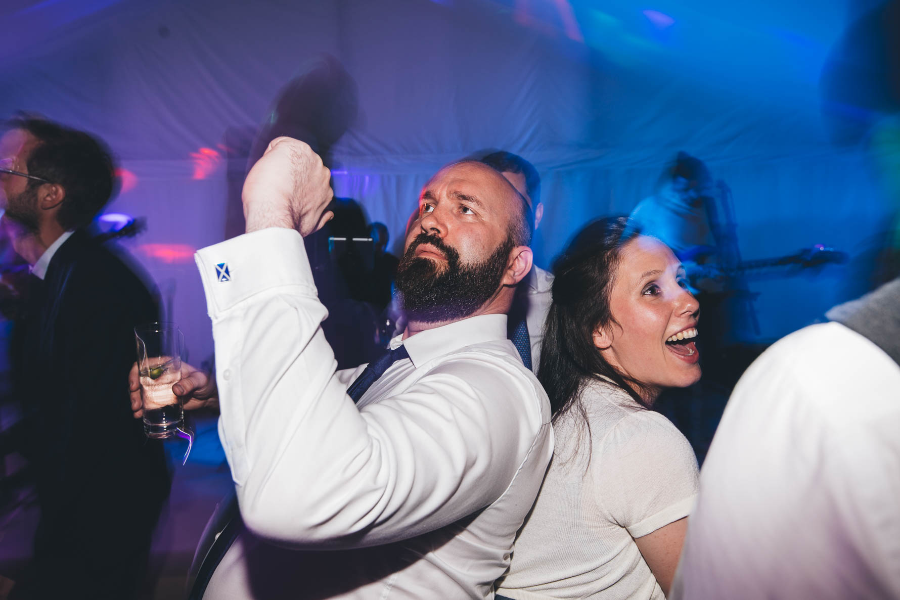 Man with a dark beard wearing a white shirt and a blue tie with his back against a woman's back as they are dancing together at a wedding reception in a marquee. She is wearing a white top. There are blue lights in the background.