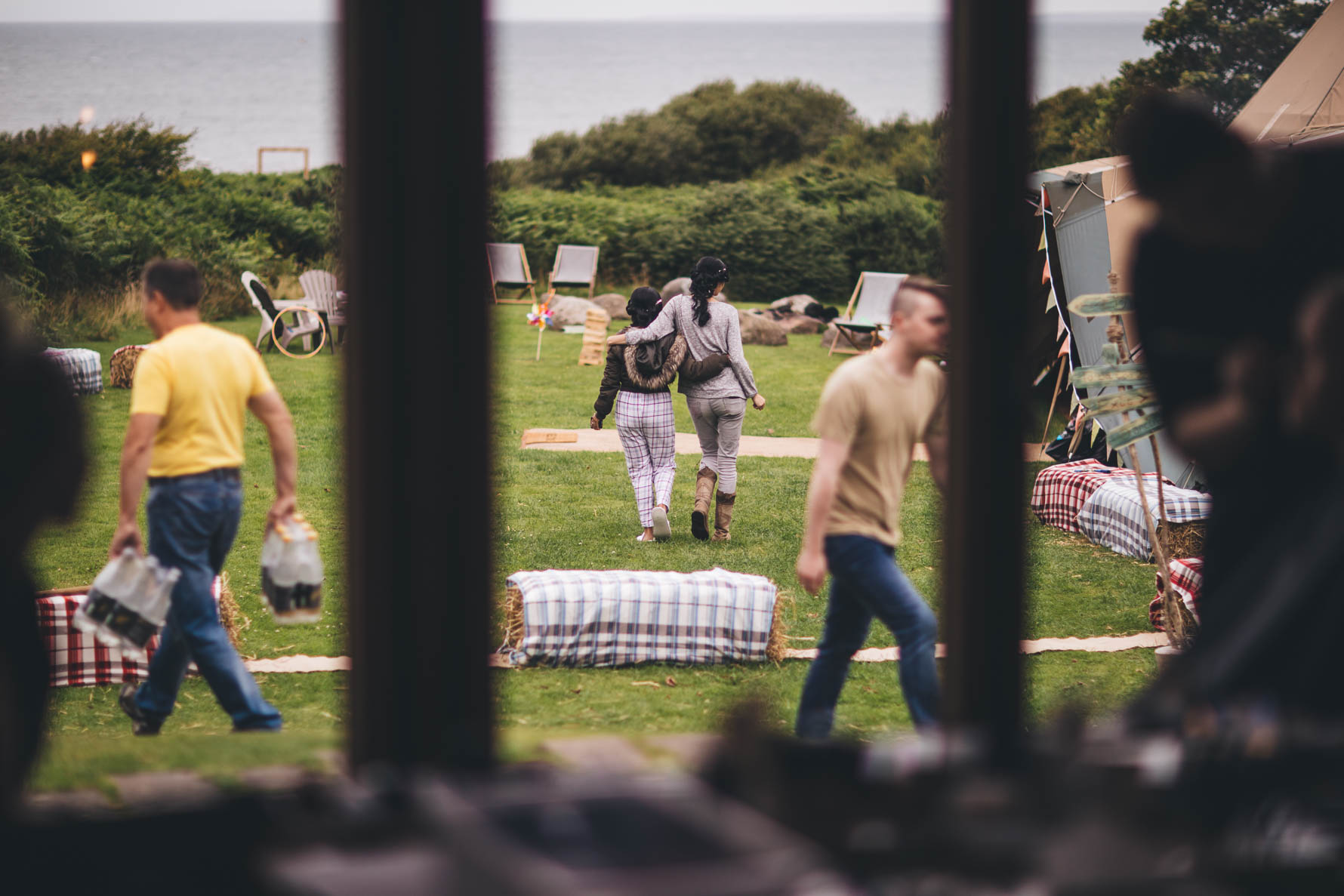 A field surrounded by bushes overlooking the sea with people busy making preparations for a wedding with hay bails covered in checked fabric for seating, deckchairs and bunting