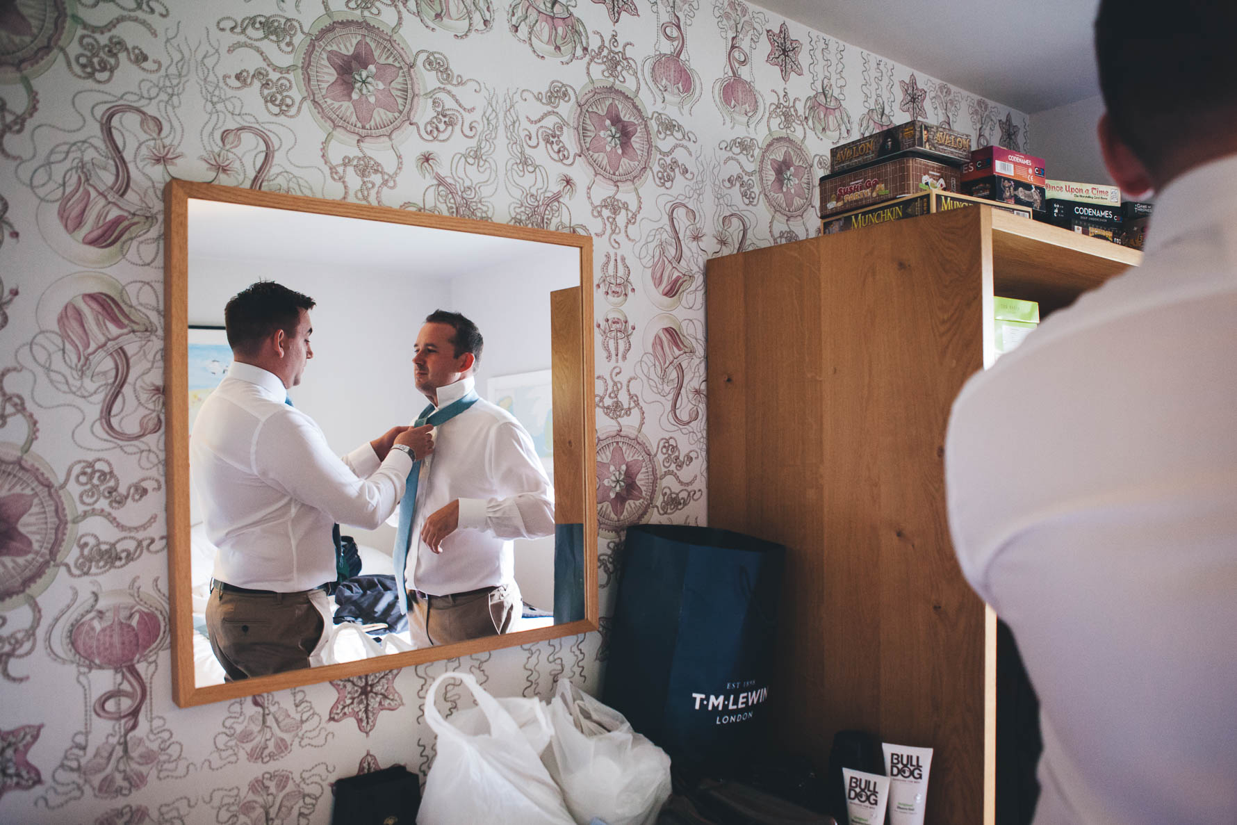 View of the mirror in a bedroom of the best man helping the groom put on his turquoise tie. Both men are wearing white shirts and chinos