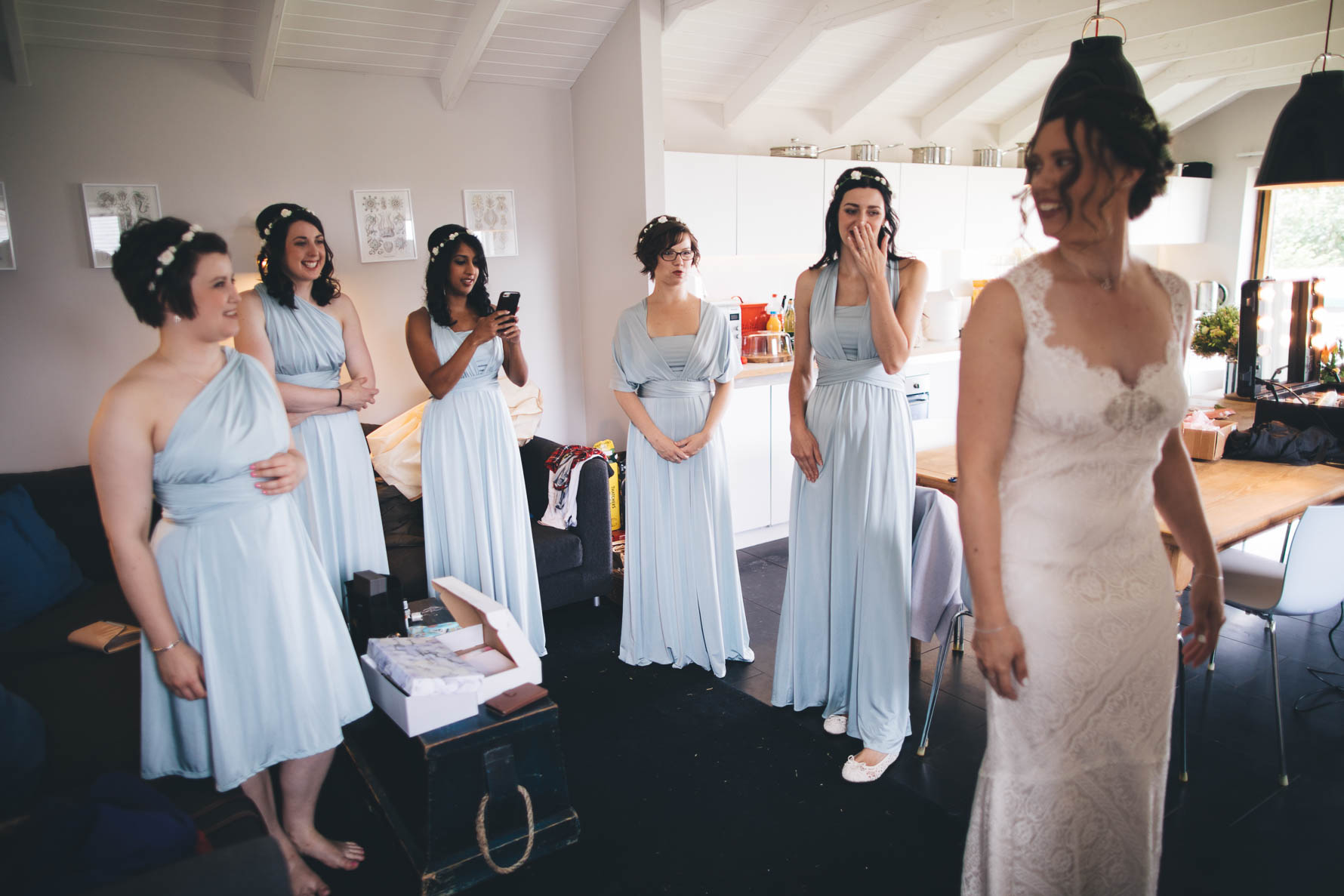 Five bridesmaids in baby blue dresses stood in a semicircle looking towards the bride who has just put her wedding dress on