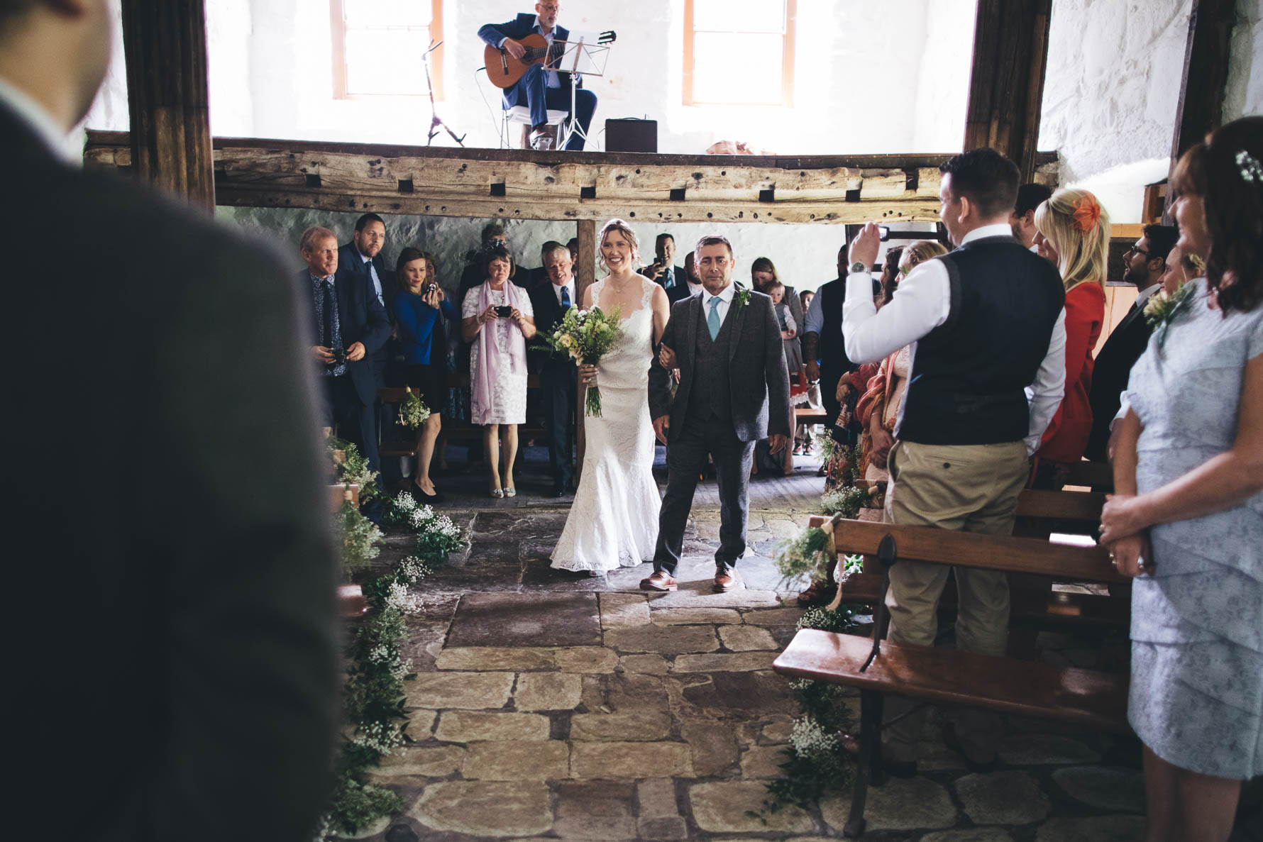 Bride and her father walking down the aisle with a man playing guitar on a platform above the aisle and wedding guests looking towards the bride