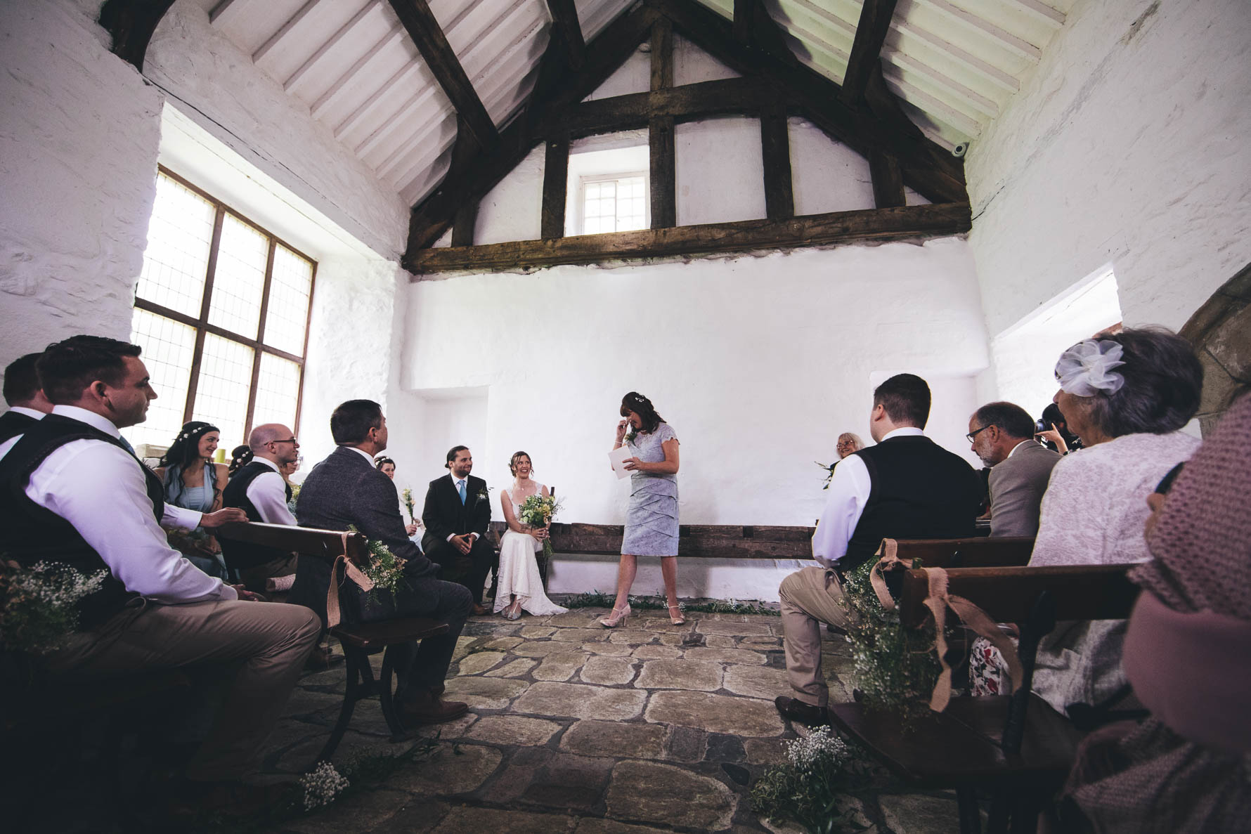 Mother of the bride stood reading a speech during the wedding ceremony in an old white washed building with wooden beams and a pitched roof whilst the rest of the wedding party are seated
