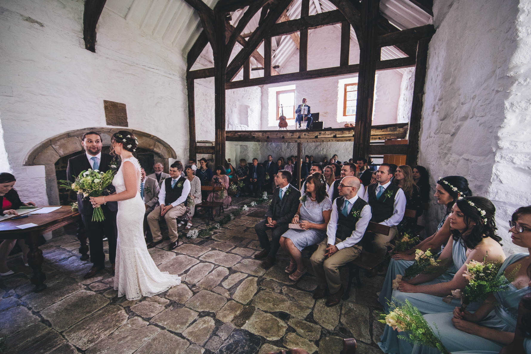 Bride holding a large bouquet of flowers stood next to the groom during their wedding ceremony in a large white washed building with wooden beams and a pitched roof