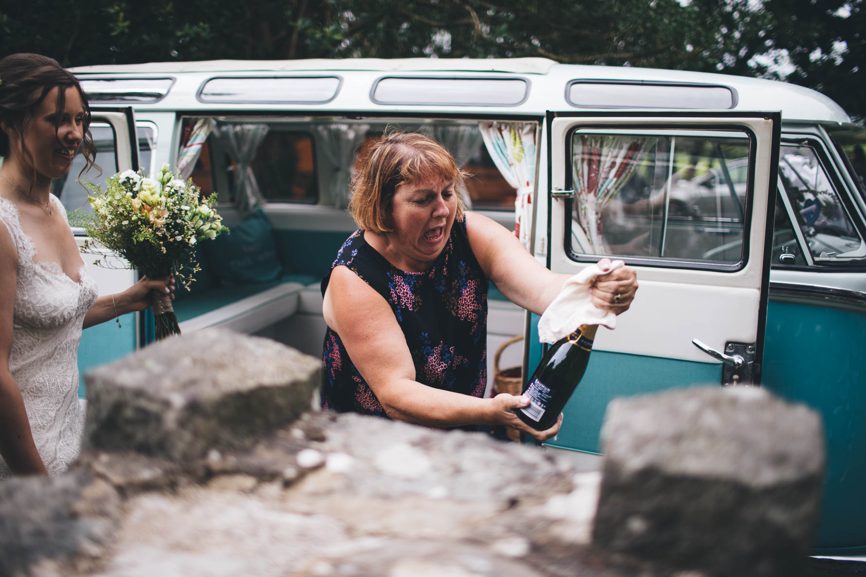 Female wedding guest opening a bottle of champagne half covered with a napkin in front of a turquoise blue vintage VW campervan as the bride looks on holding a bouquet of flowers