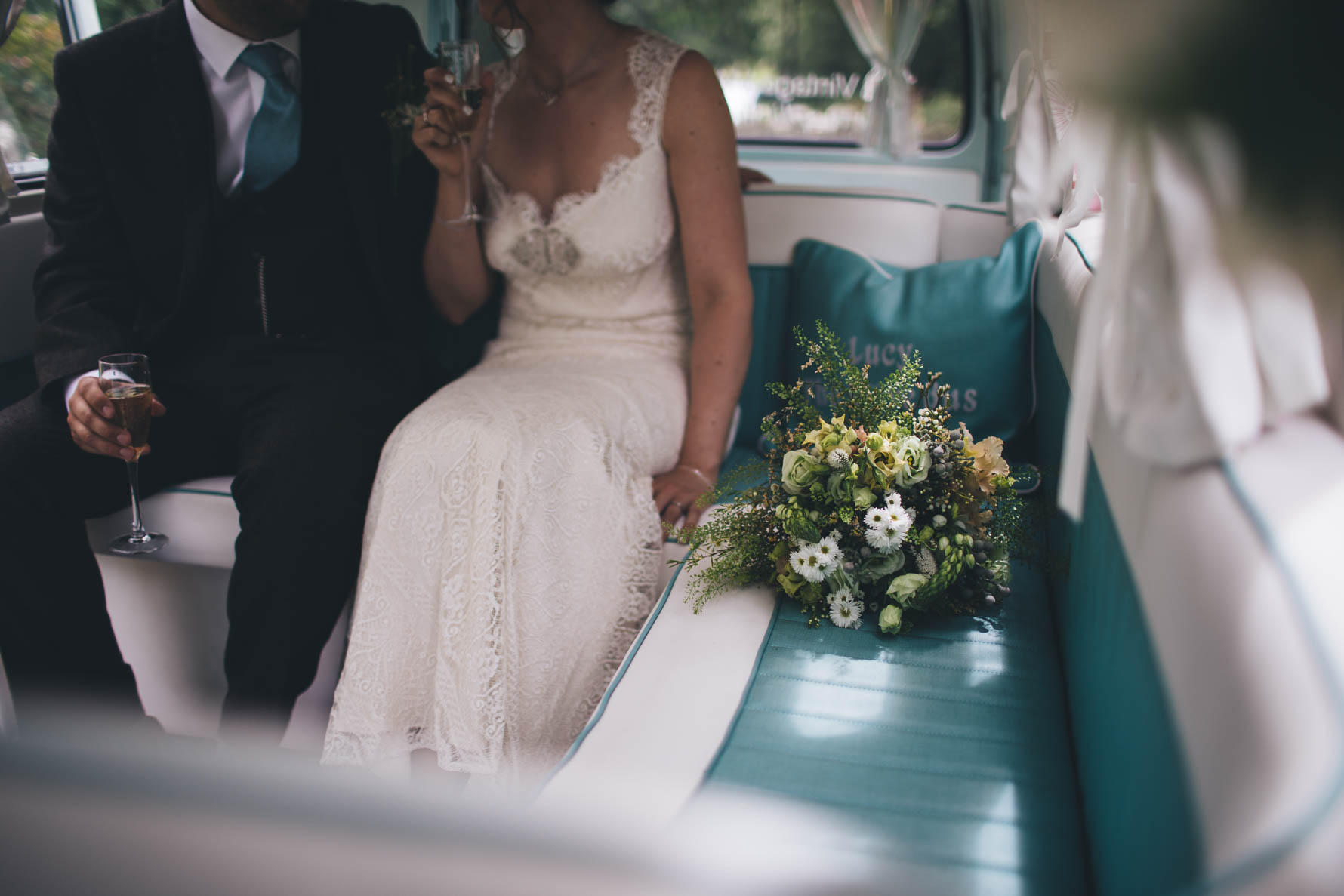 Bride and groom sat in a vintage VW campervan holding glasses of champagne with a bouquet of flowers sat on the seat beside the bride. You can only see the bride and groom from the neck down
