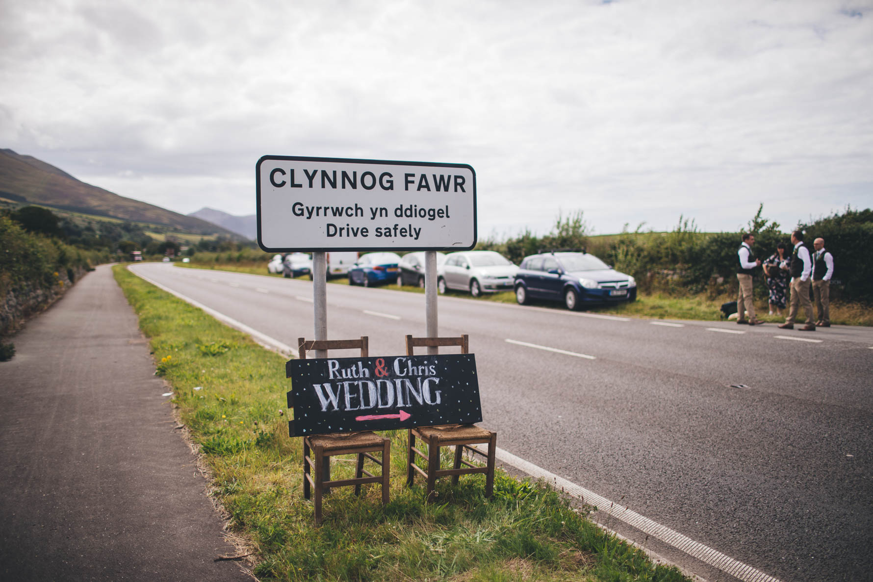 Road sign with a chalk board below announcing Ruth & Chris' wedding with an arrow pointing to the right which is sat on two chairs. There are cars parked up on the right ha=nd side of the road and four wedding guests stood in front of the cars