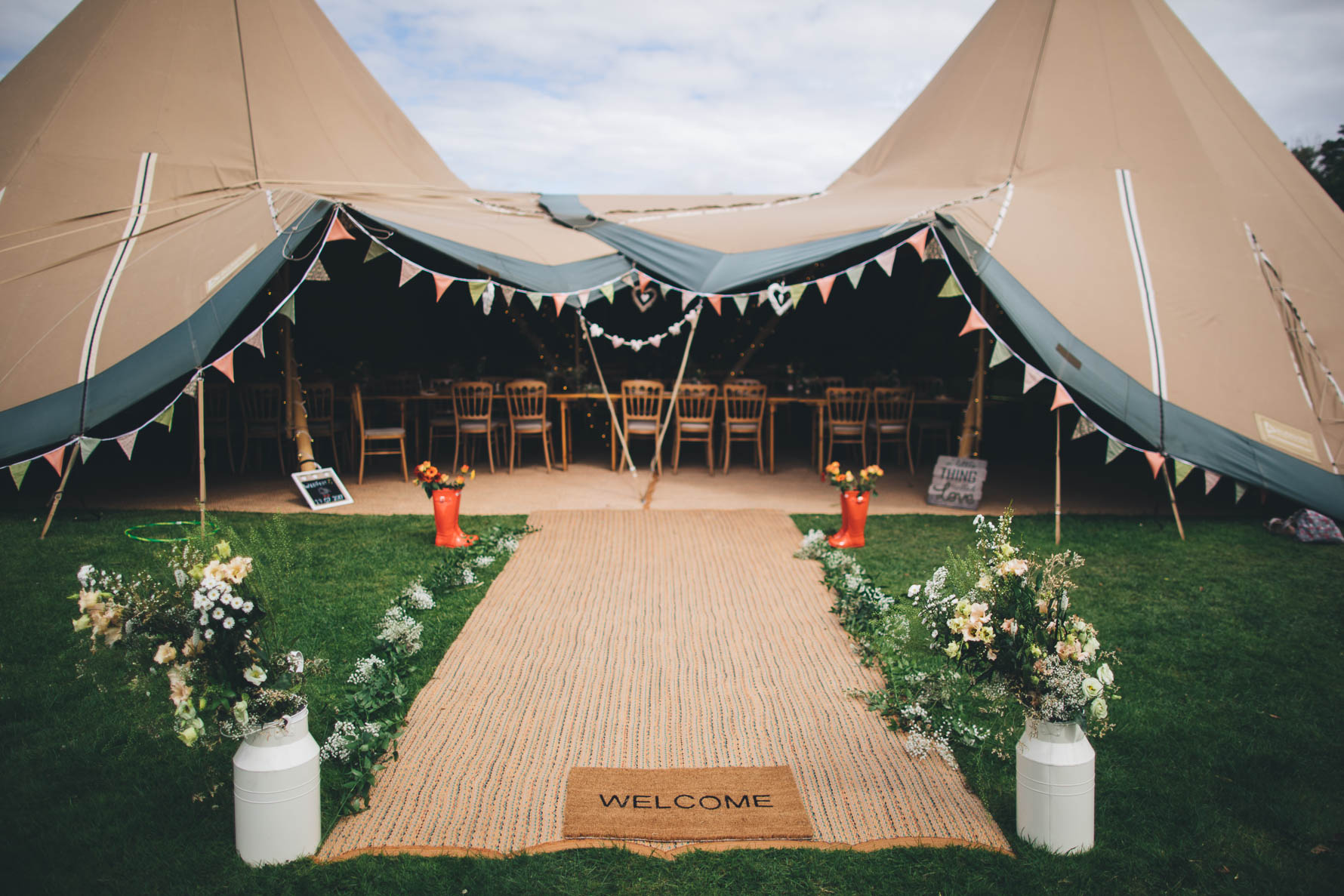 Entrance to a tipi set up for a wedding reception with a welcome mat and flowers at the entrance and bunting around the opening with tables and chairs inside