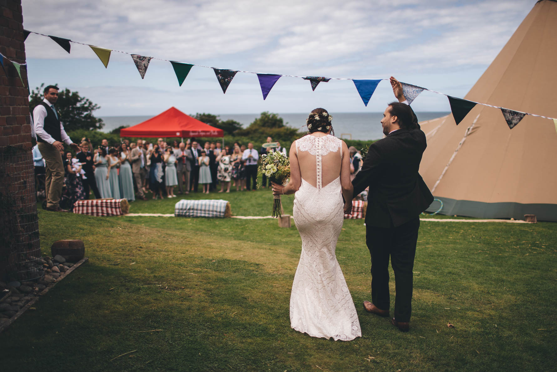 Bride and groom walking towards their wedding guests under a line of bunting which the groom is holding up to allow the couple to walk underneath. The bride and groom are walking away from the camera. There is a tipi to the right hand side of the shot