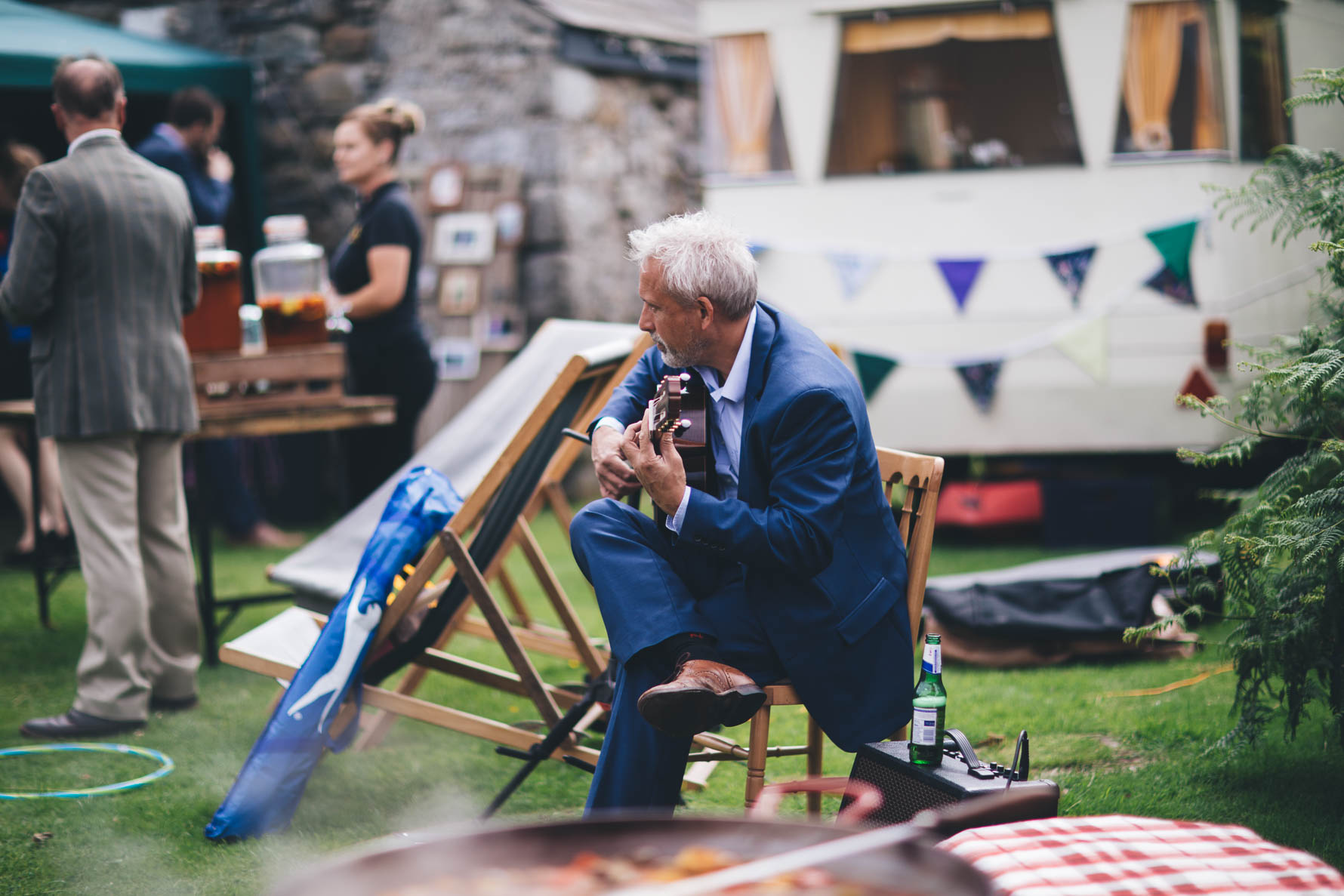 Man with grey hair and a blue suit sat on a chair outside playing a guitar