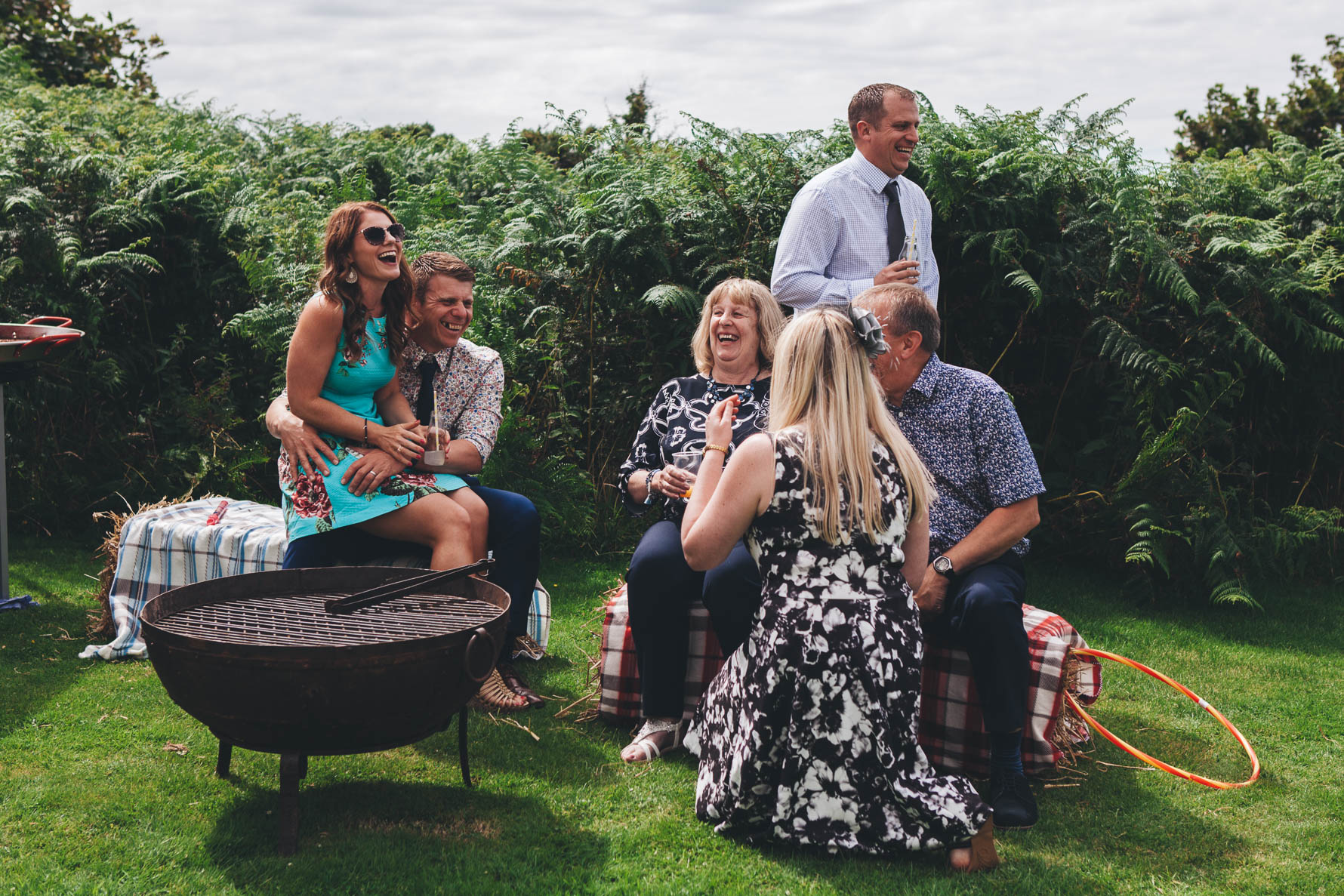 Wedding guests sat on bails of hay in front of an unlit metal barbeque. All of the guests are laughing