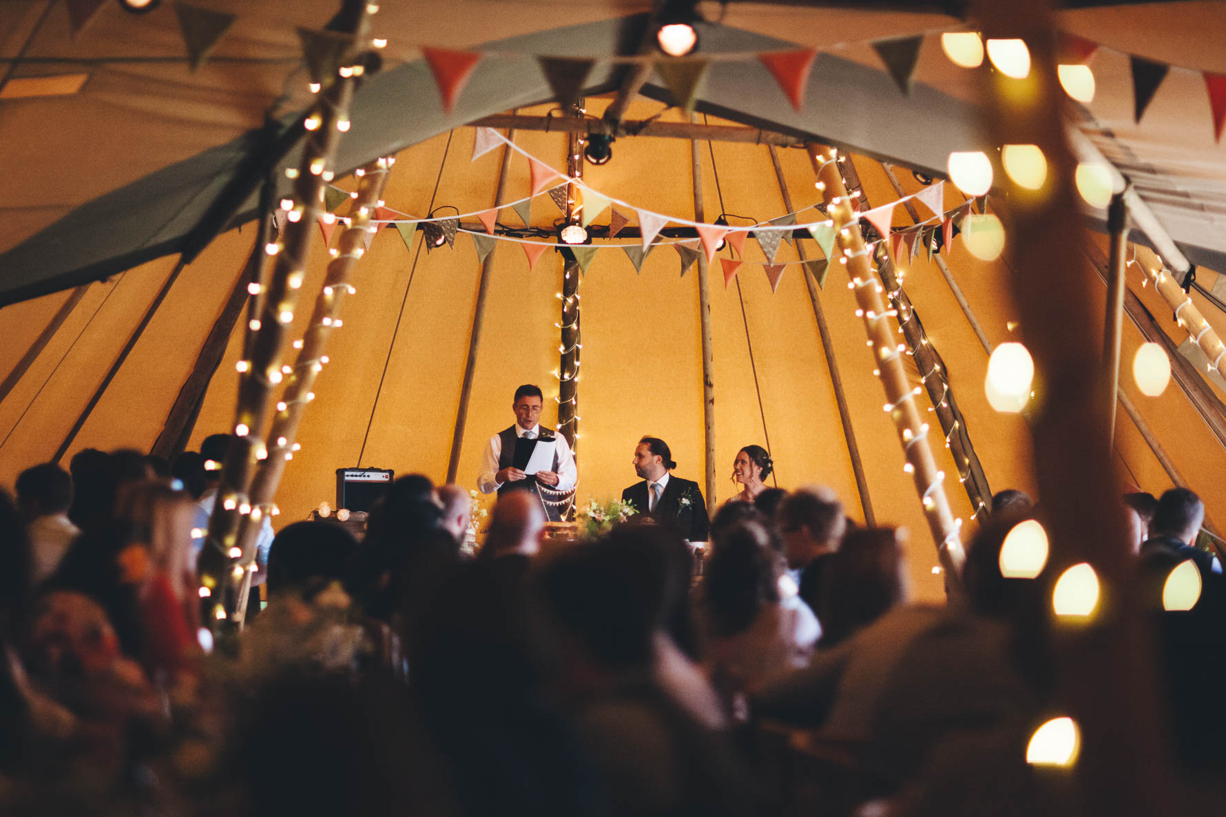 Best man stood reading a speech inside a tipi with bunting and fairy lights around the supports of the tipi