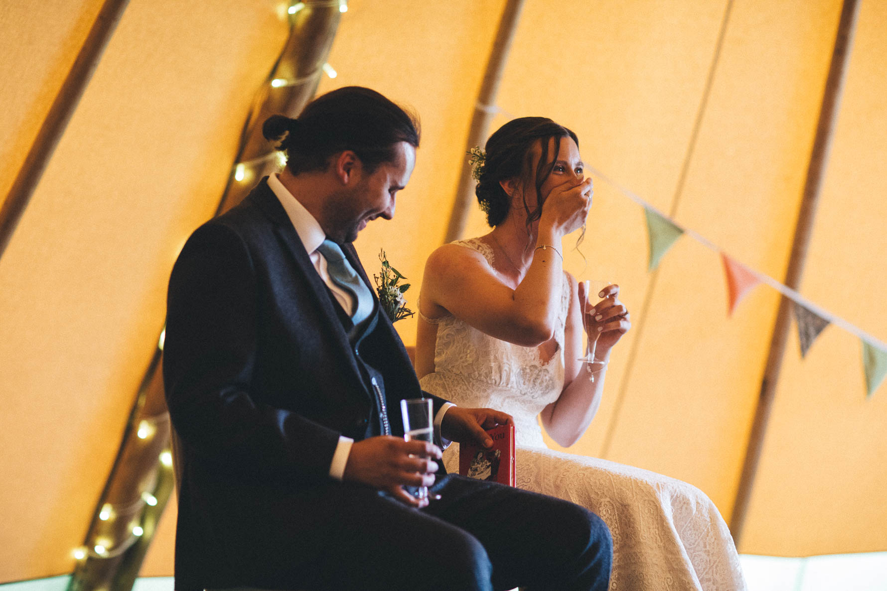 Bride and groom sat laughing as a speech is being read out. There are fairylights and bunting behind them against the side of the yellow tipi they are sat in