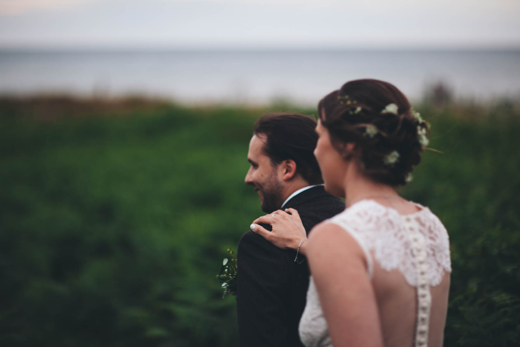 Bride walking with her hand on the groom's shoulder as they walk through a filed which is out of focus. You can see the sea in the distance