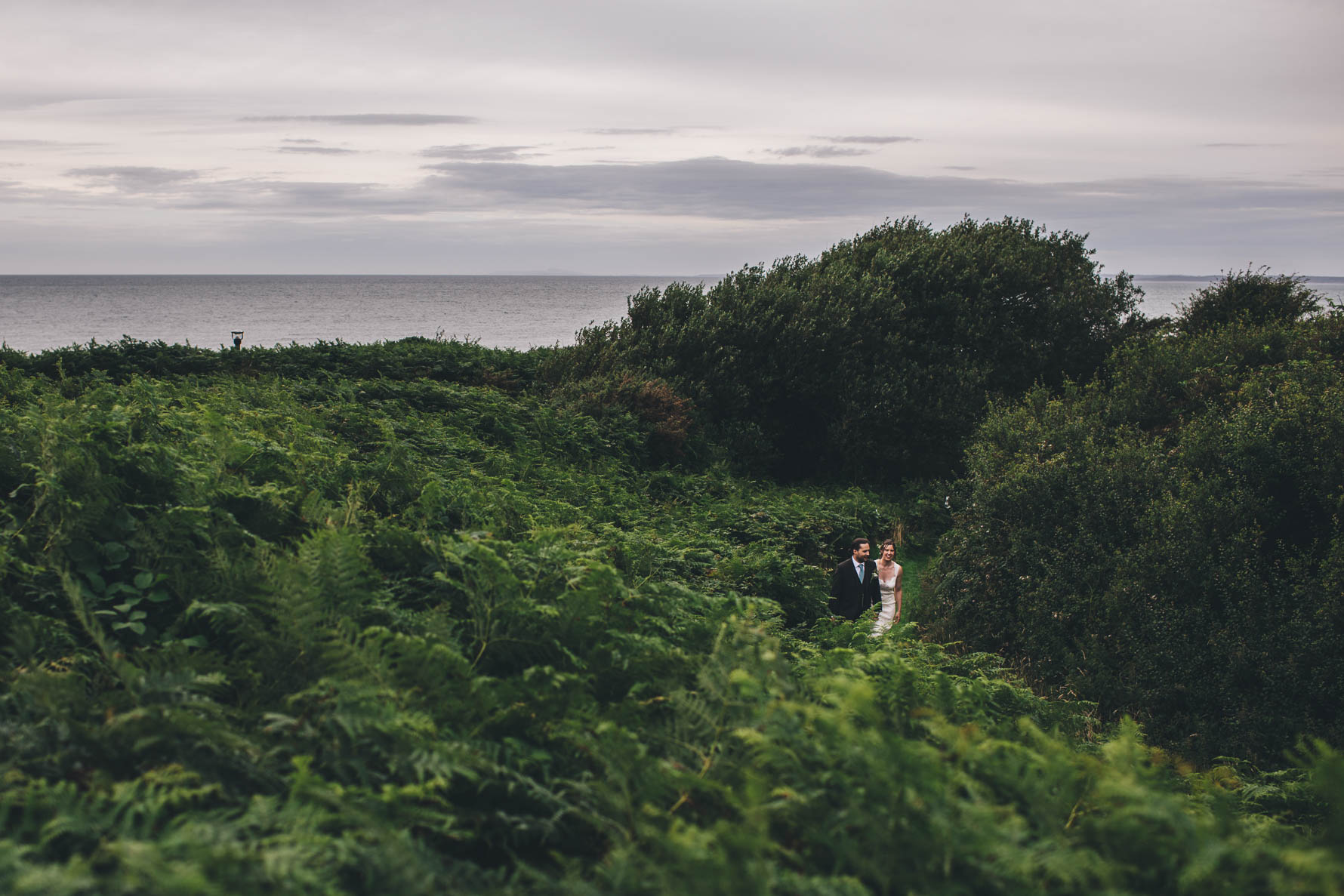 Bride and groom walking along a path amongst lots of tall ferns and trees by the sea
