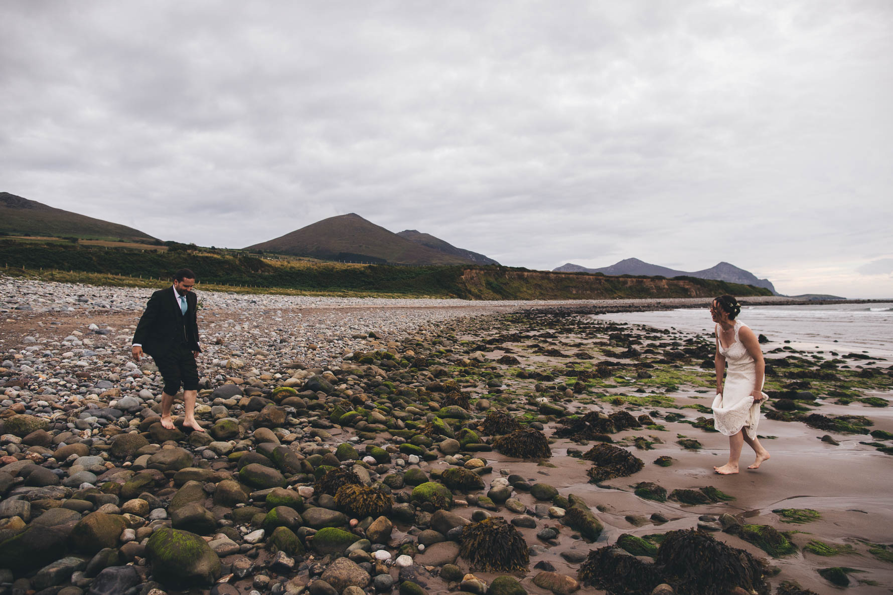 Bride and groom barefoot as they walk over pebbles an rocks at the seaside with mountains in the background