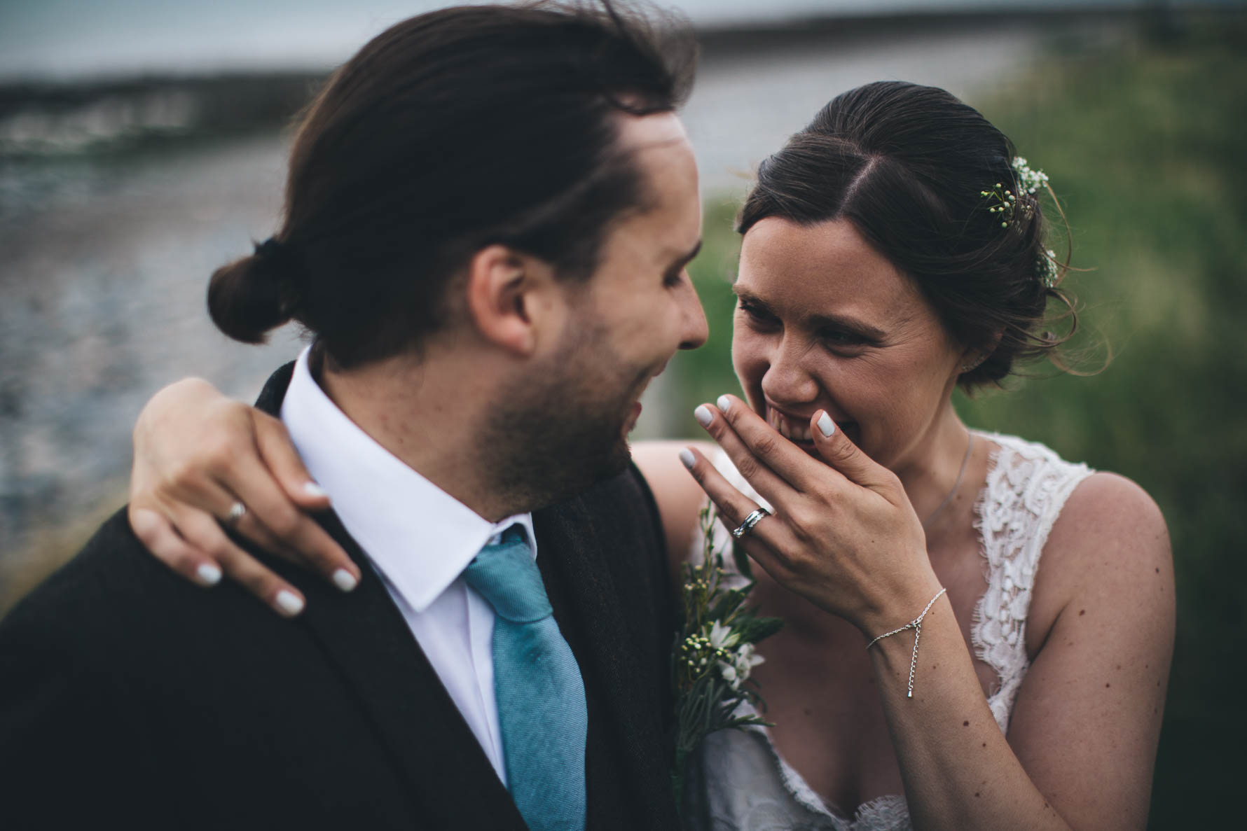 Bride has one hand around the groom's neck and her other covering her mouth as she is laughing as she looks towards the groom